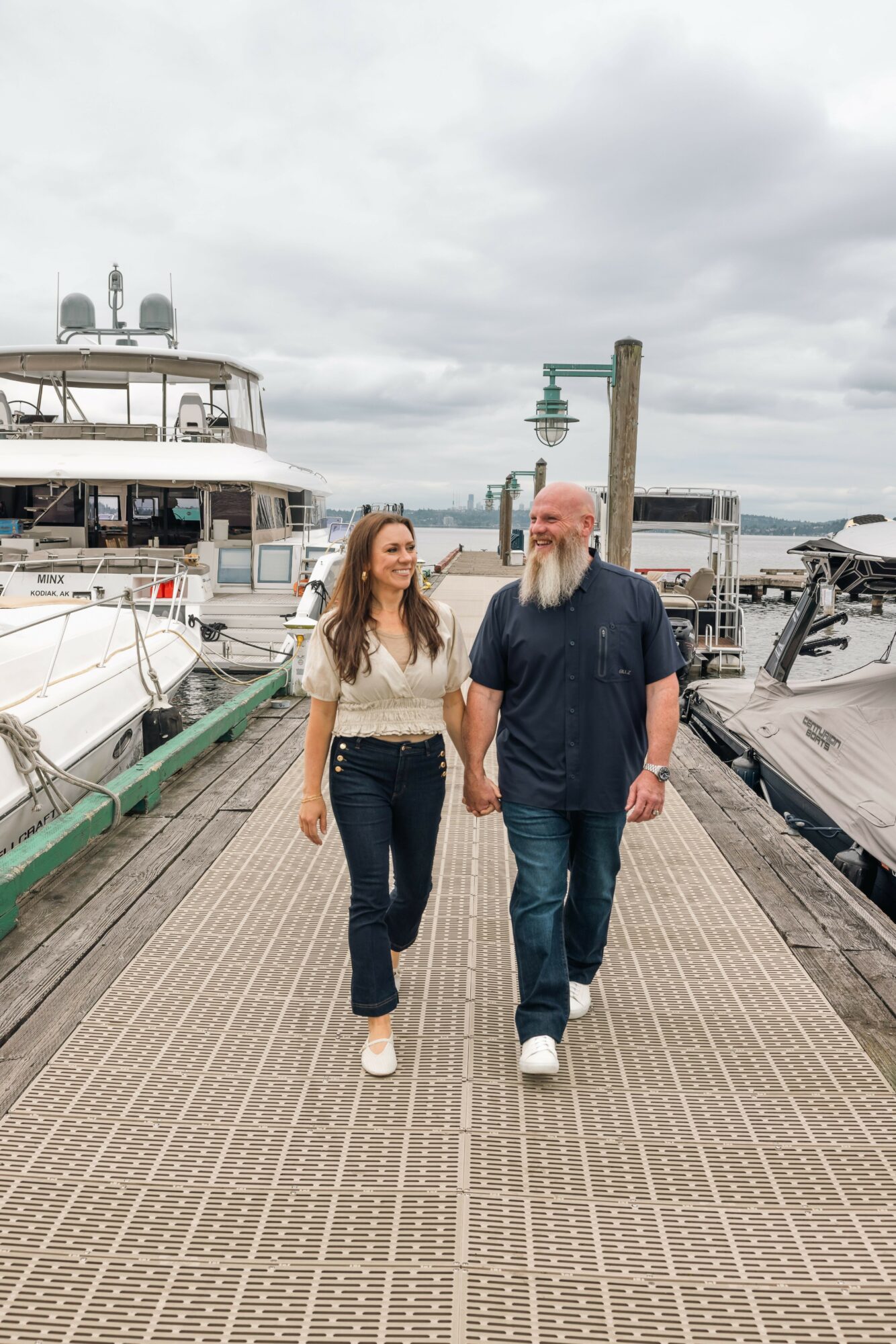 A woman and a man walk hand in hand on a dock with boats and yachts in the background under cloudy sky.