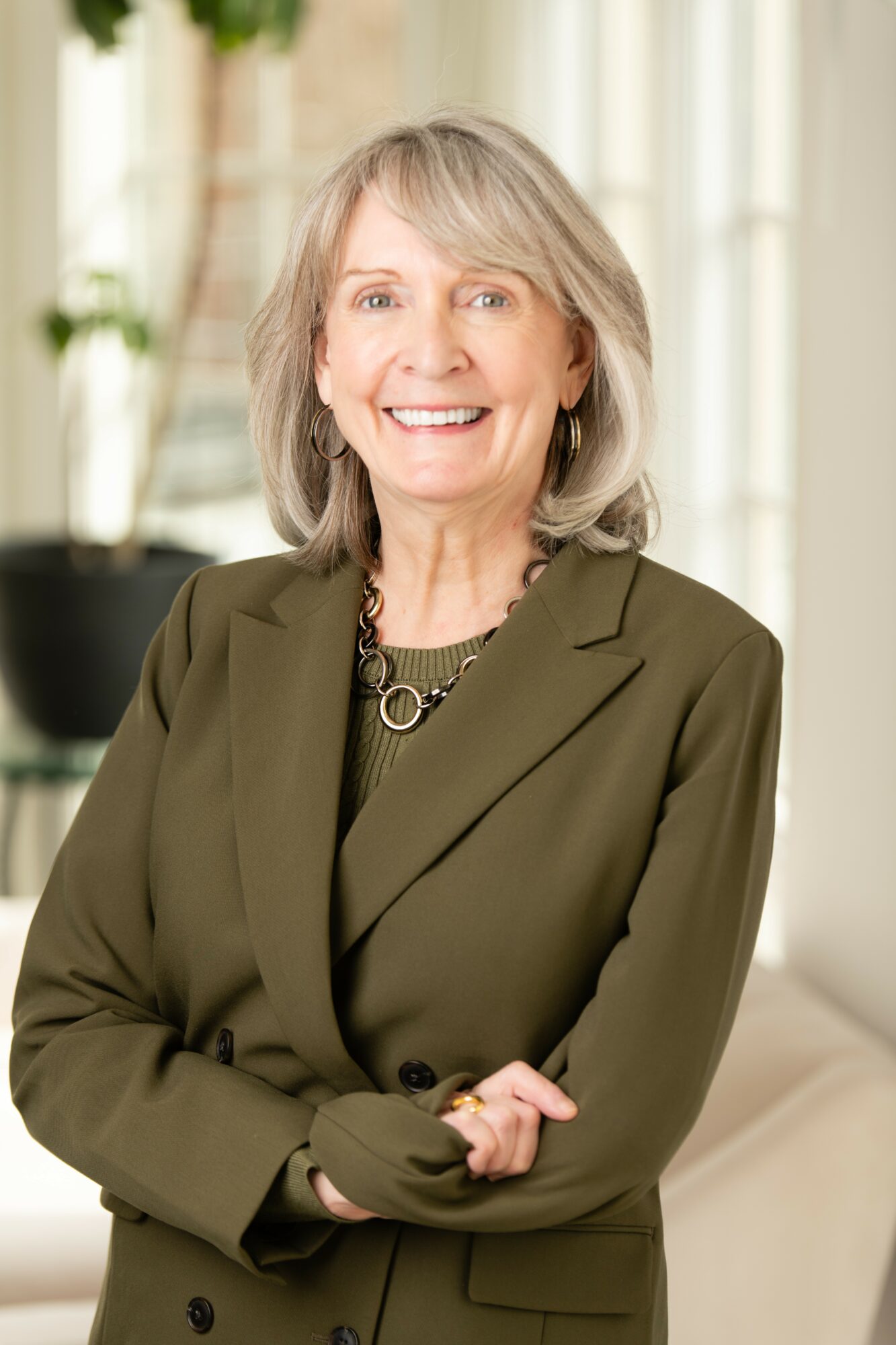 Smiling woman with shoulder-length gray hair wearing a green blazer and jewelry, standing indoors with a blurred background.
