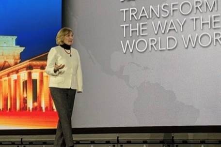 Woman standing on stage with a large screen behind her displaying text and an image of a building, speaking to an audience.