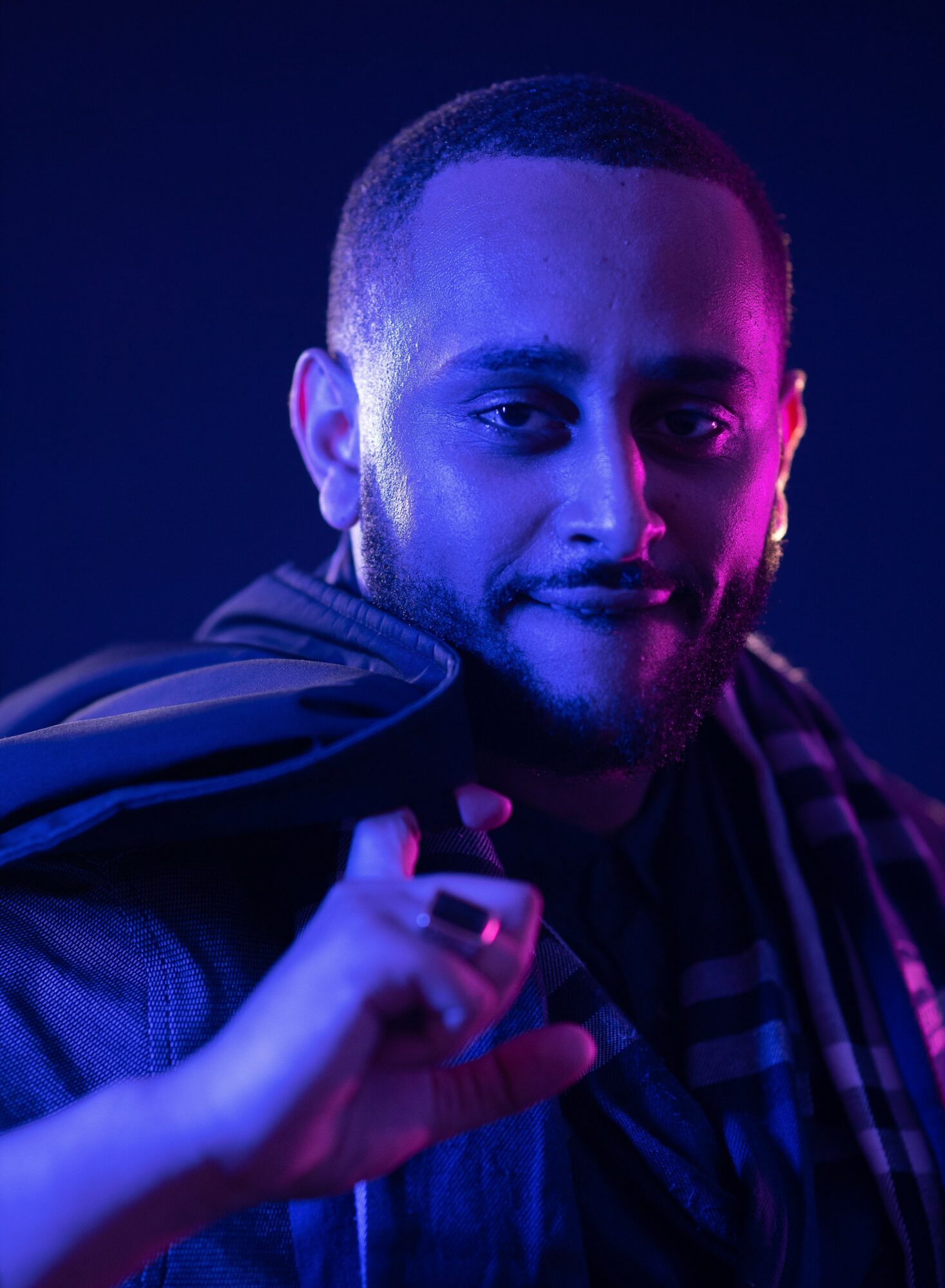 Portrait of a man with short hair, smiling slightly, illuminated by blue and purple lighting, wearing a jacket and scarf.
