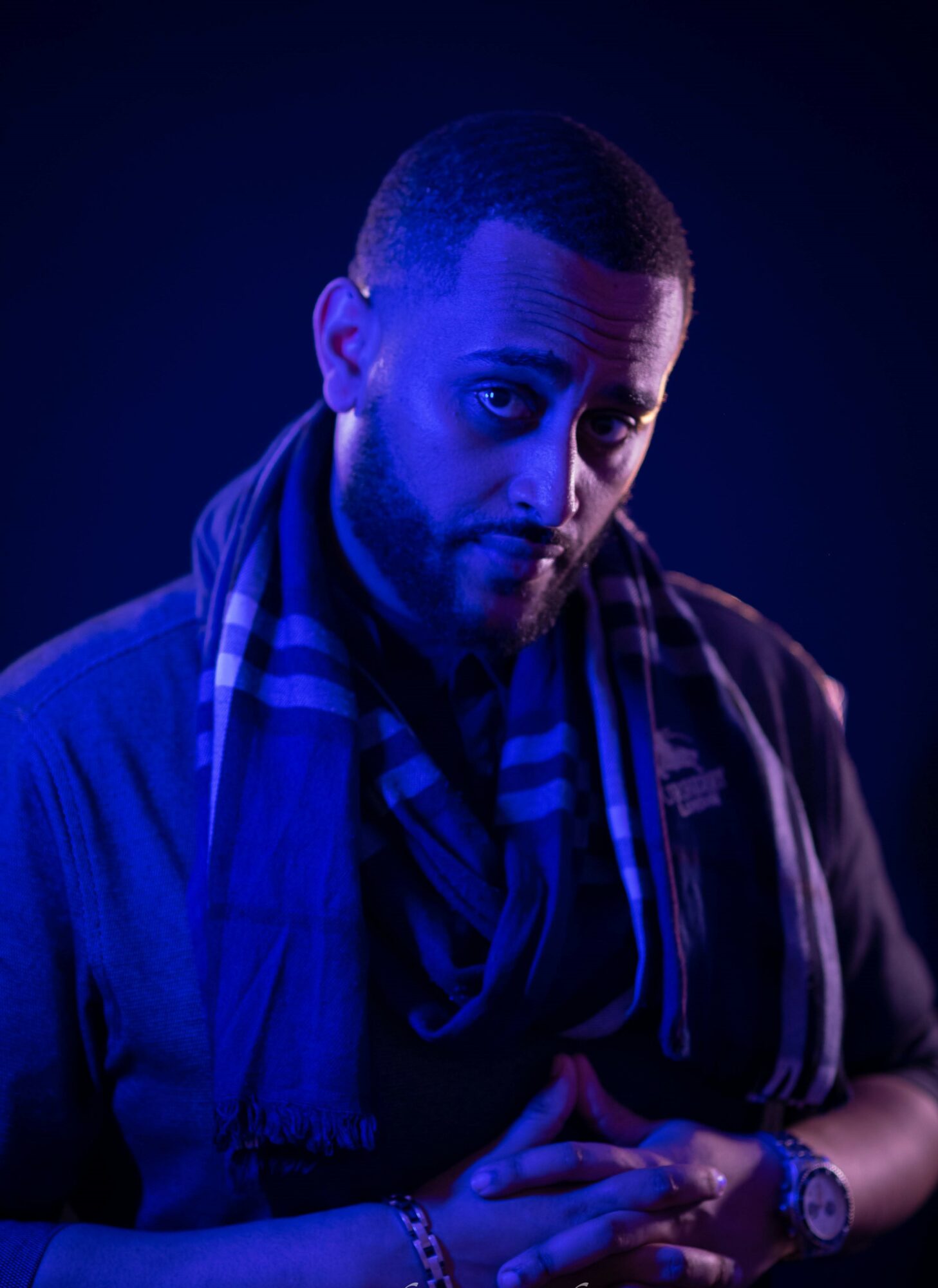 Man with short hair and beard wearing a dark jacket and scarf, looking at camera with hands clasped, dark background