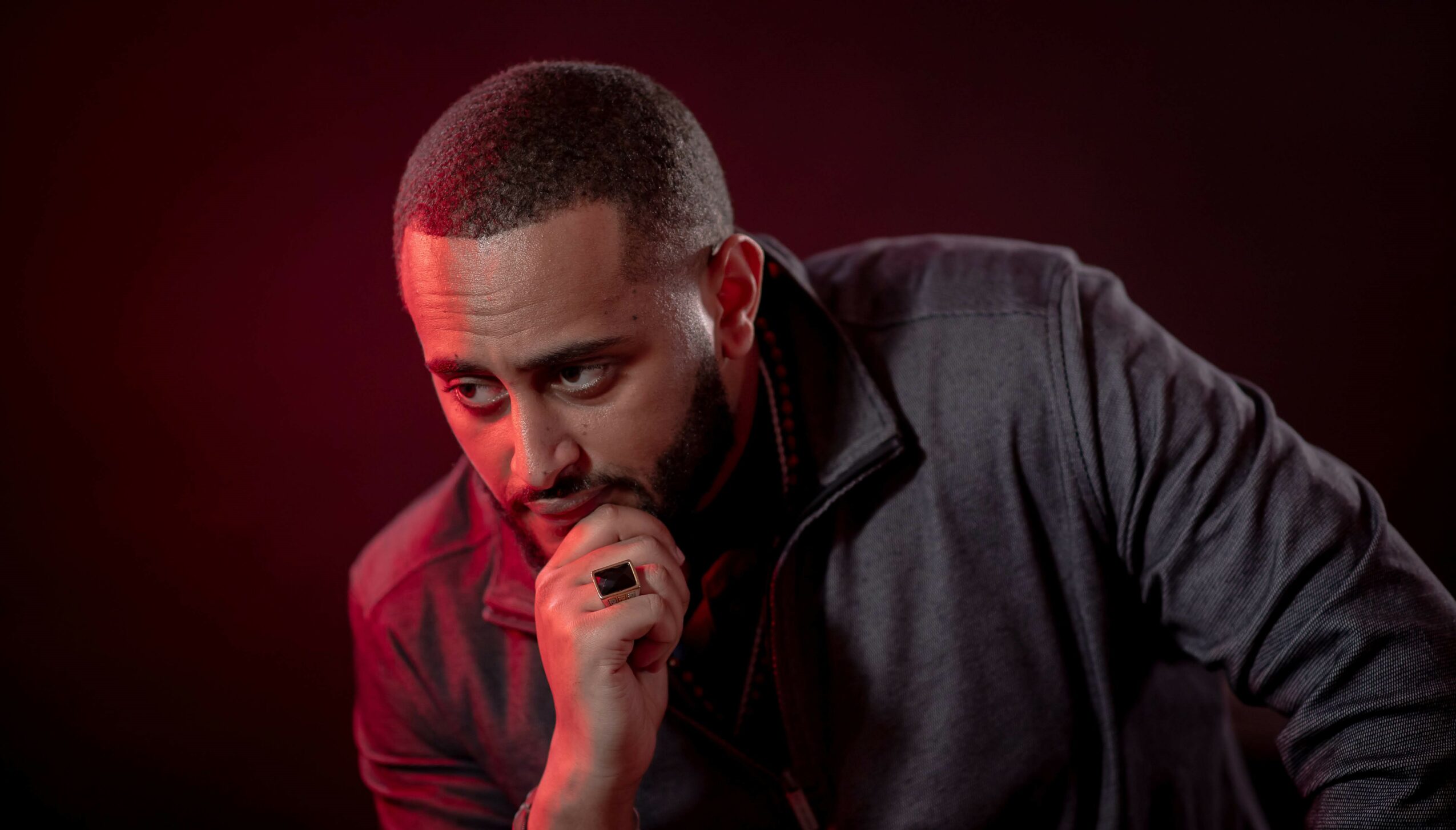 Man with short hair and beard, wearing a dark jacket, looking thoughtful against a dark background with red lighting.