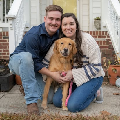 A smiling couple with a golden retriever sitting on steps outside a house, holding the dog between them.