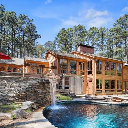 Wooden house with large windows, surrounded by trees, with a pool and waterfall feature in the foreground.