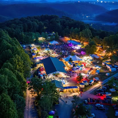 Night scene of an outdoor festival with tents, colorful lights, and people, surrounded by trees and hills.