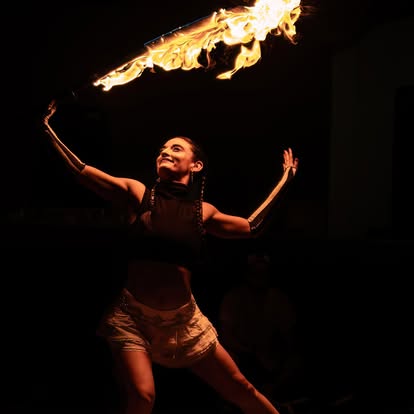 Person performing fire breathing stunt with flames in dark background, smiling, arms raised, wearing shorts and a top.