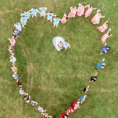 People form a heart shape on grass, with a person in the center holding a child, viewed from above.