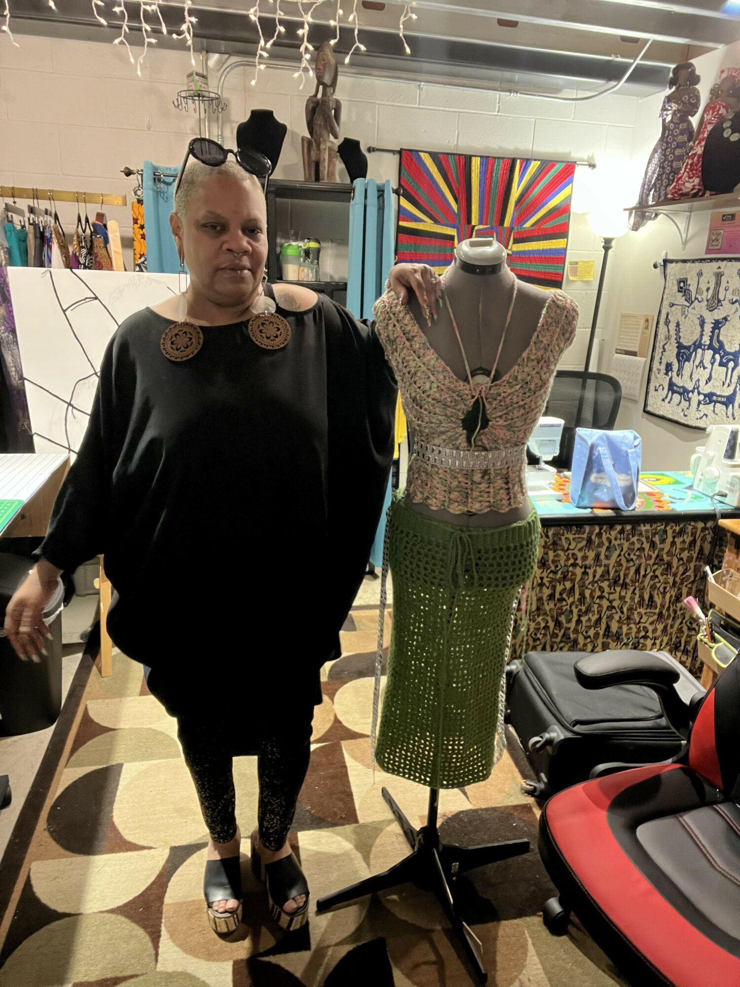 Woman standing next to a mannequin dressed in a lace top and green skirt in a boutique, with clothing and accessories in the background.