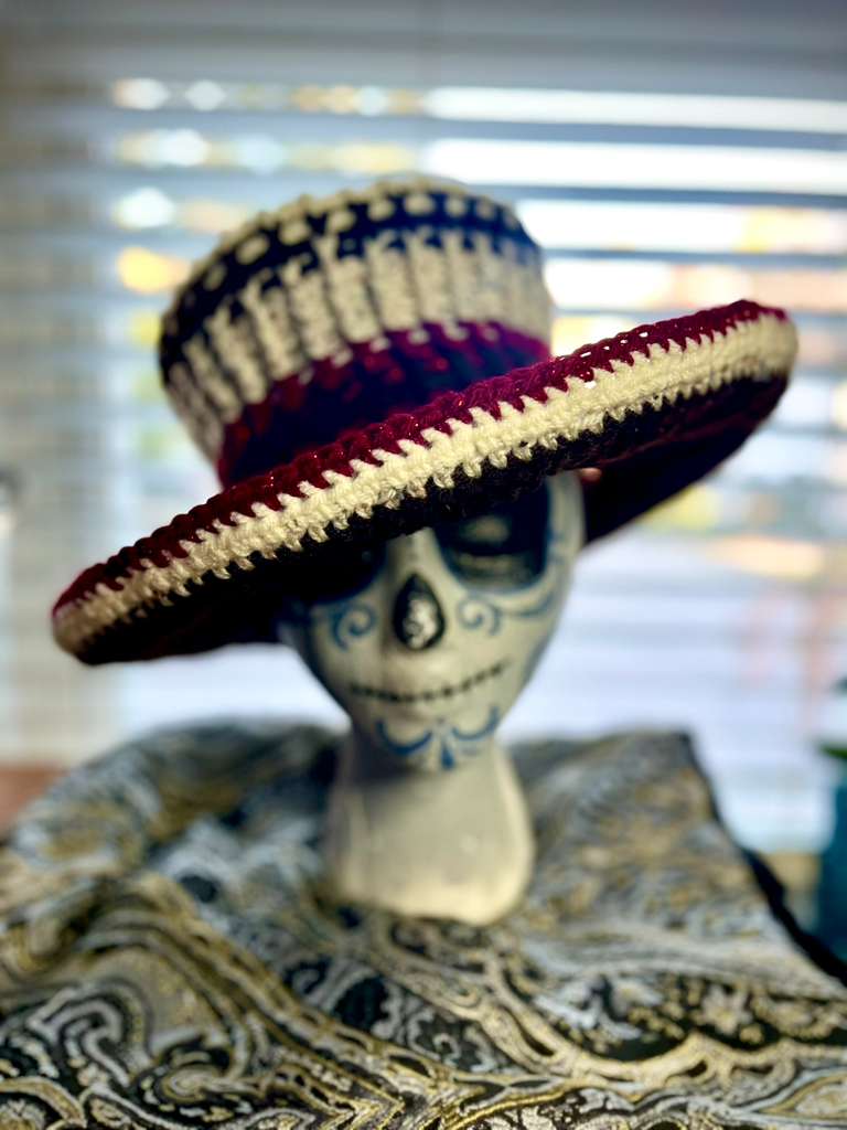 Decorative skull with a colorful sombrero, placed on patterned fabric, with blinds in the background.