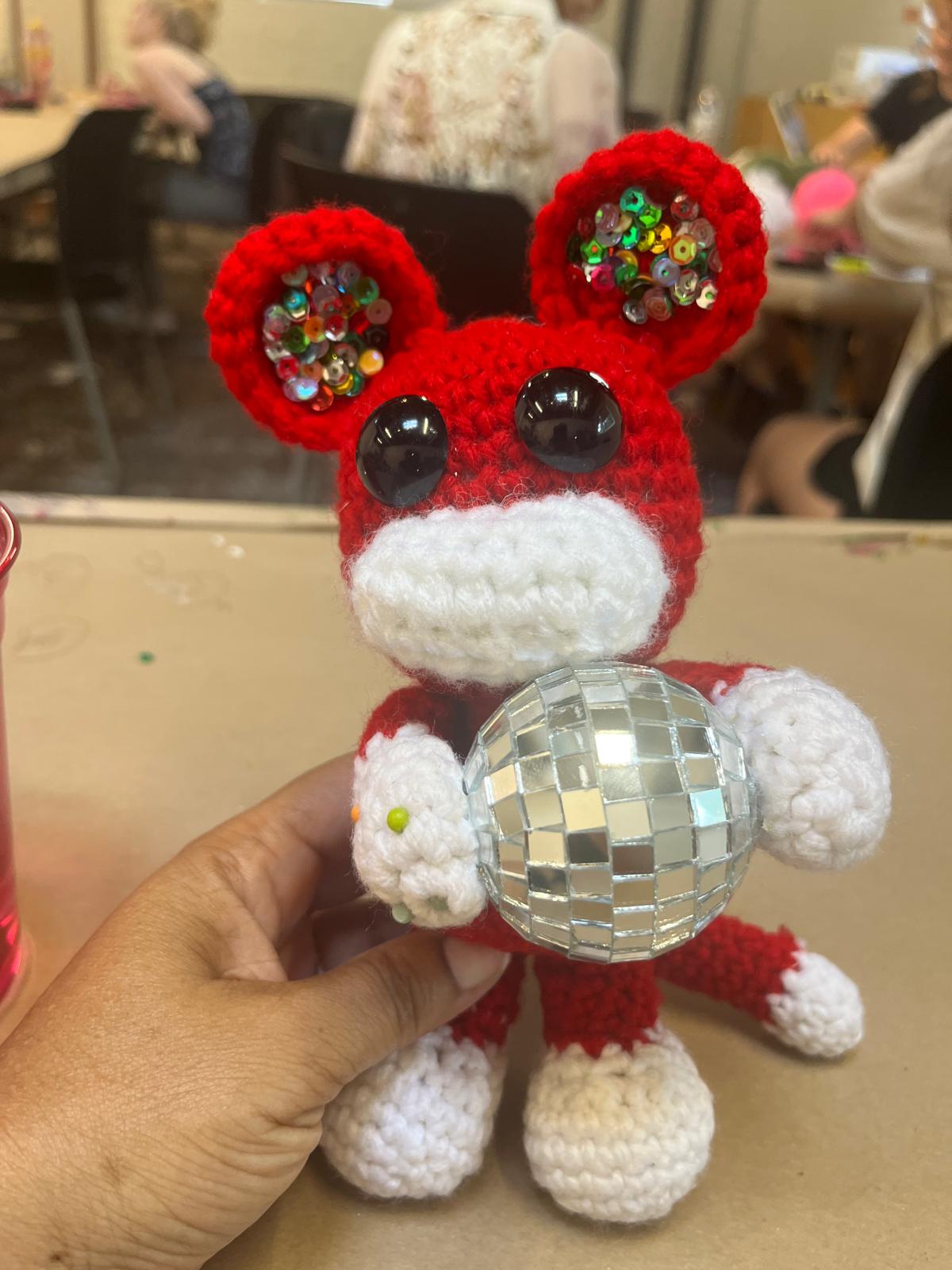 Hand holding a small crocheted mouse with red ears, black eyes, and a disco ball belly, in a room with people in the background.