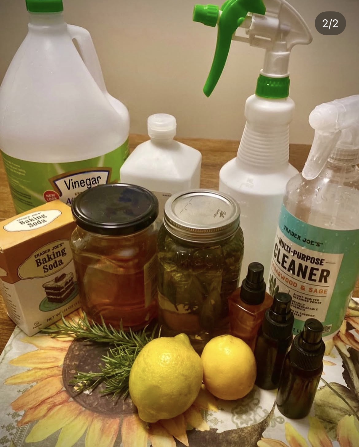 Various cleaning supplies, jars with herbs, two lemons, and small bottles on a floral tablecloth.