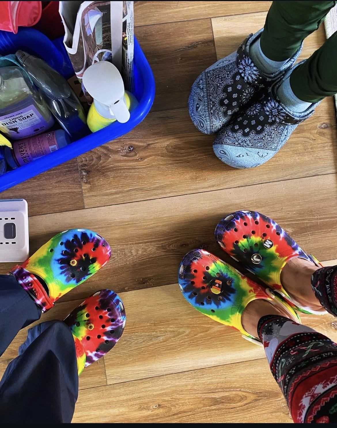Four people wearing colorful tie-dye shoes standing on a wooden floor, with a blue basket and a pair of patterned slippers nearby.