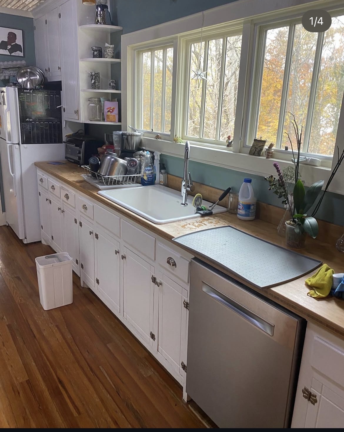 Kitchen with white cabinets, wooden countertop, large window, sink, and various appliances, plants, and items on the counter.