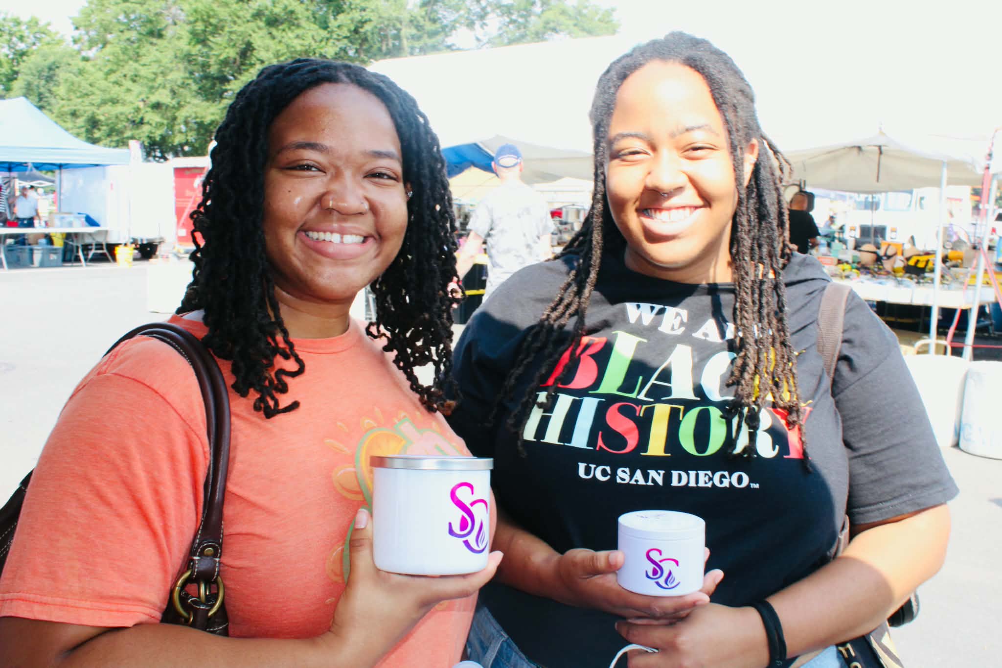 Two smiling women holding small white cups outdoors at a festival or fair, with tents and trees in the background.