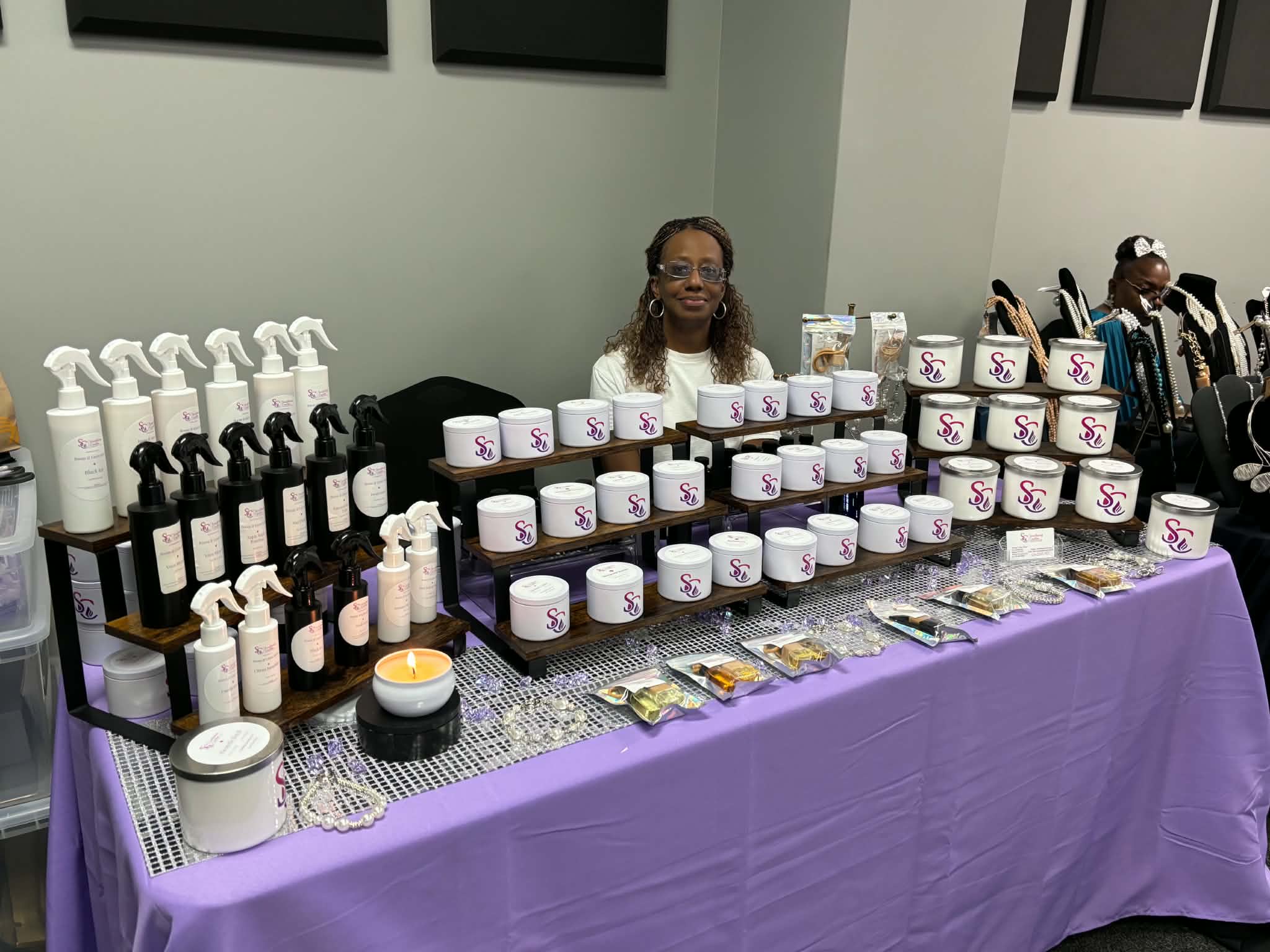 Woman sitting behind a table with skincare products, jars, bottles, and samples, covered with a purple tablecloth, in an indoor setting.