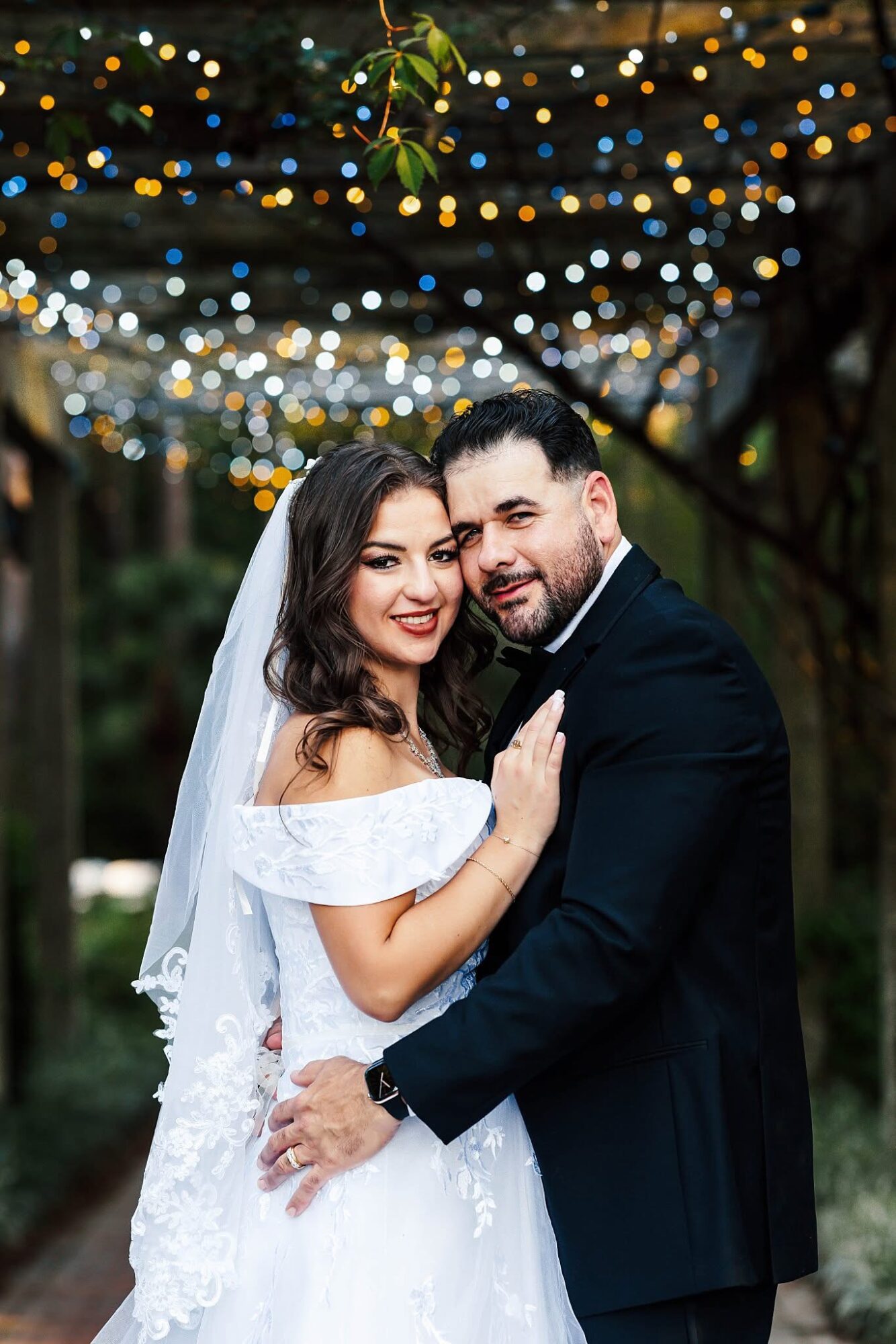 Bride and groom smiling outdoors with string lights in background, bride in white dress and veil, groom in dark suit.