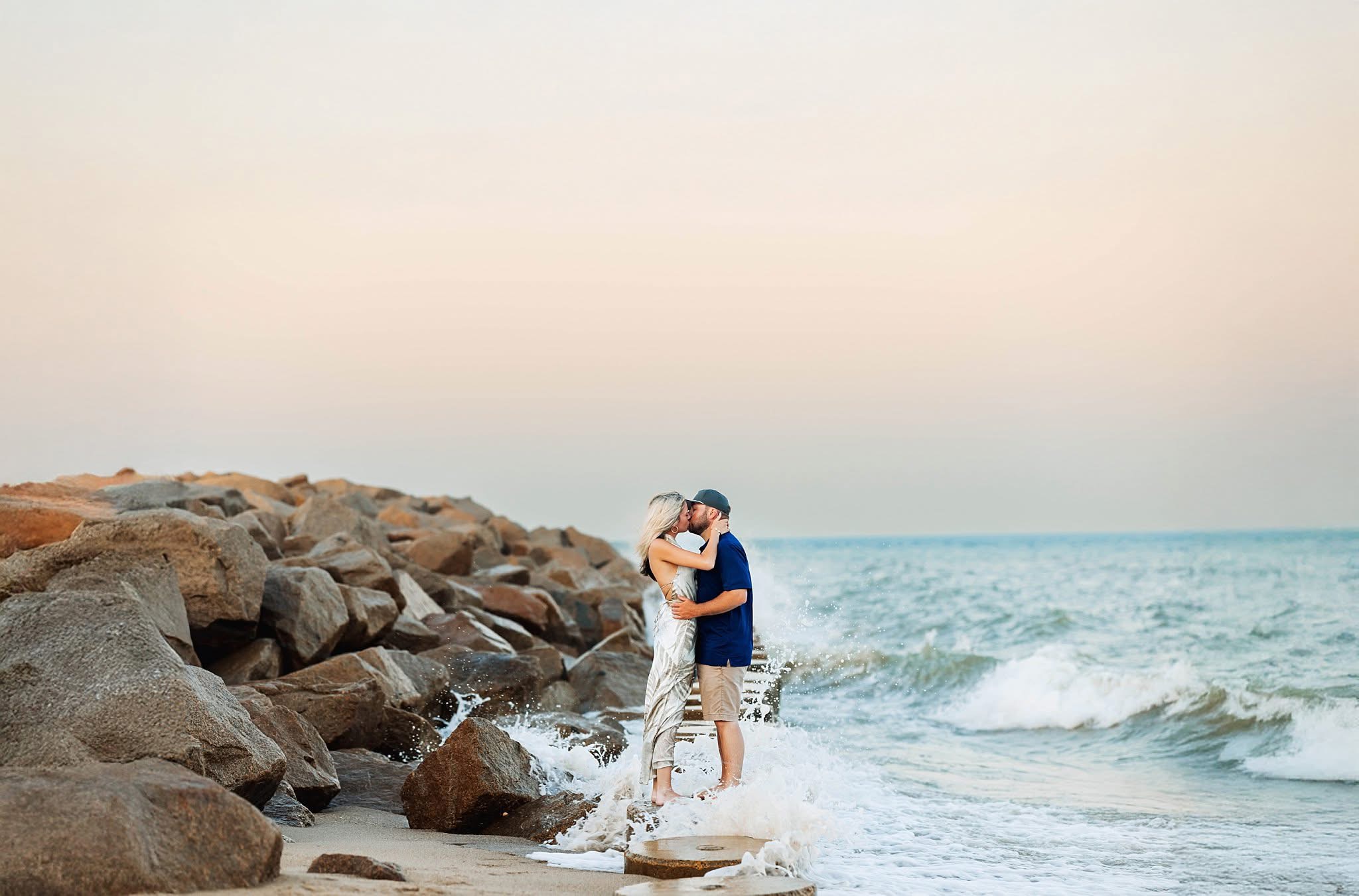 Two people hugging on a rocky beach with ocean waves and a clear sky in the background.