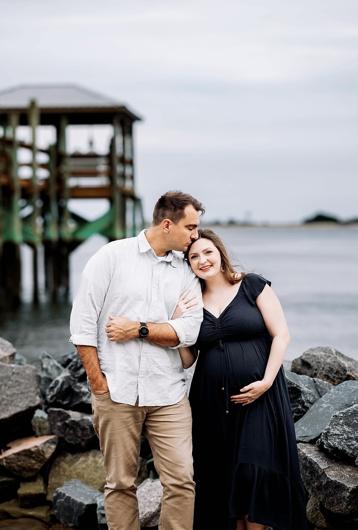 A couple stands on rocks near water, with a pier in the background, smiling and embracing, woman pregnant, man wearing a white shirt.