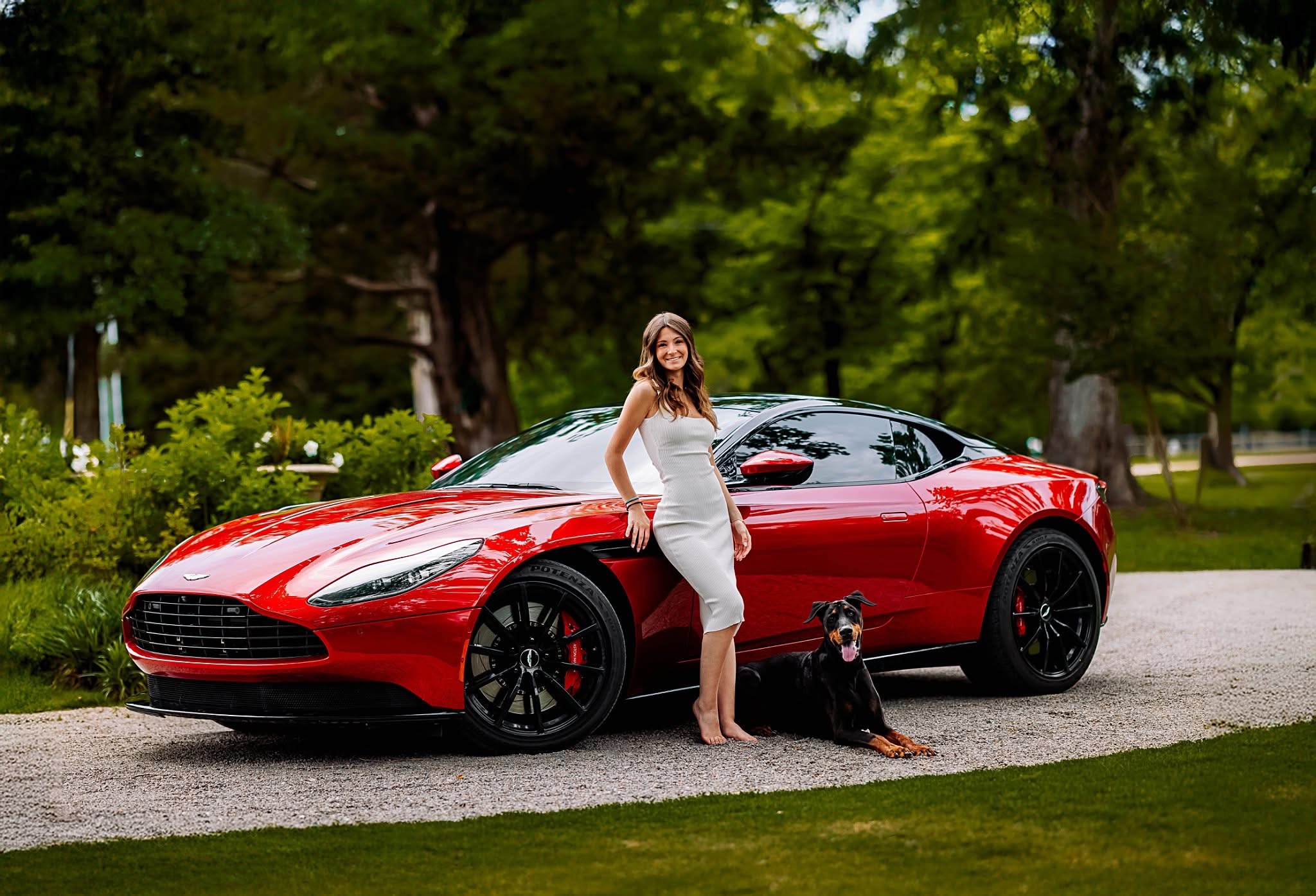 Woman in white dress standing next to a red sports car with a dog sitting nearby, outdoors with trees in background.