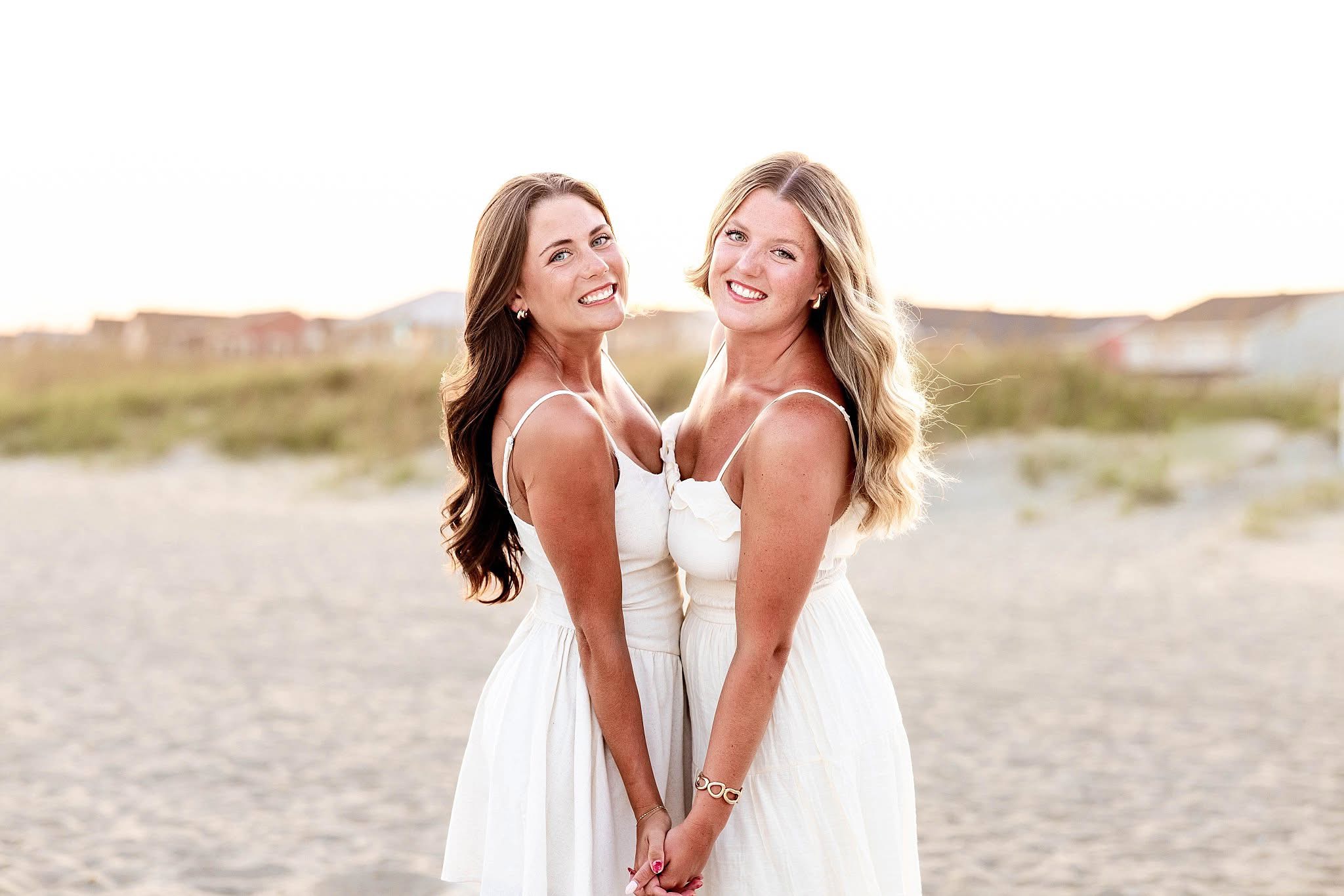 Two women in white dresses standing outdoors on a sunny day, smiling and holding hands, with a blurred landscape background.