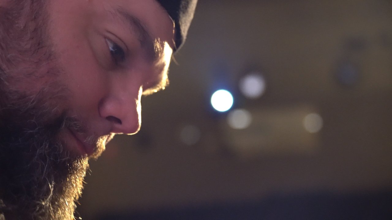 Close-up of a person's face with a beard, looking downward, with blurred background lights.