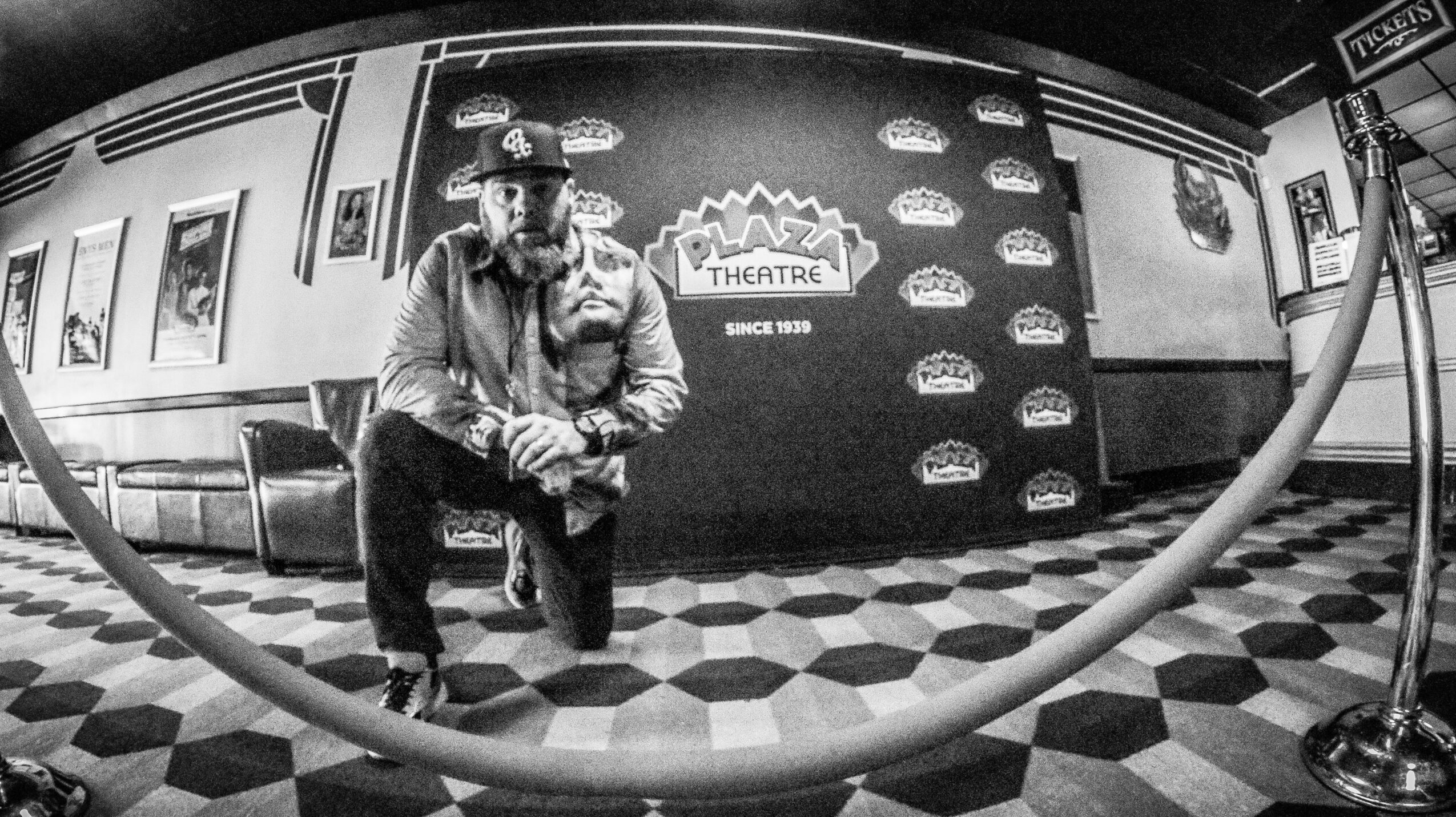 Person crouching in front of a theater backdrop with a patterned floor and a rope barrier, black and white photo.