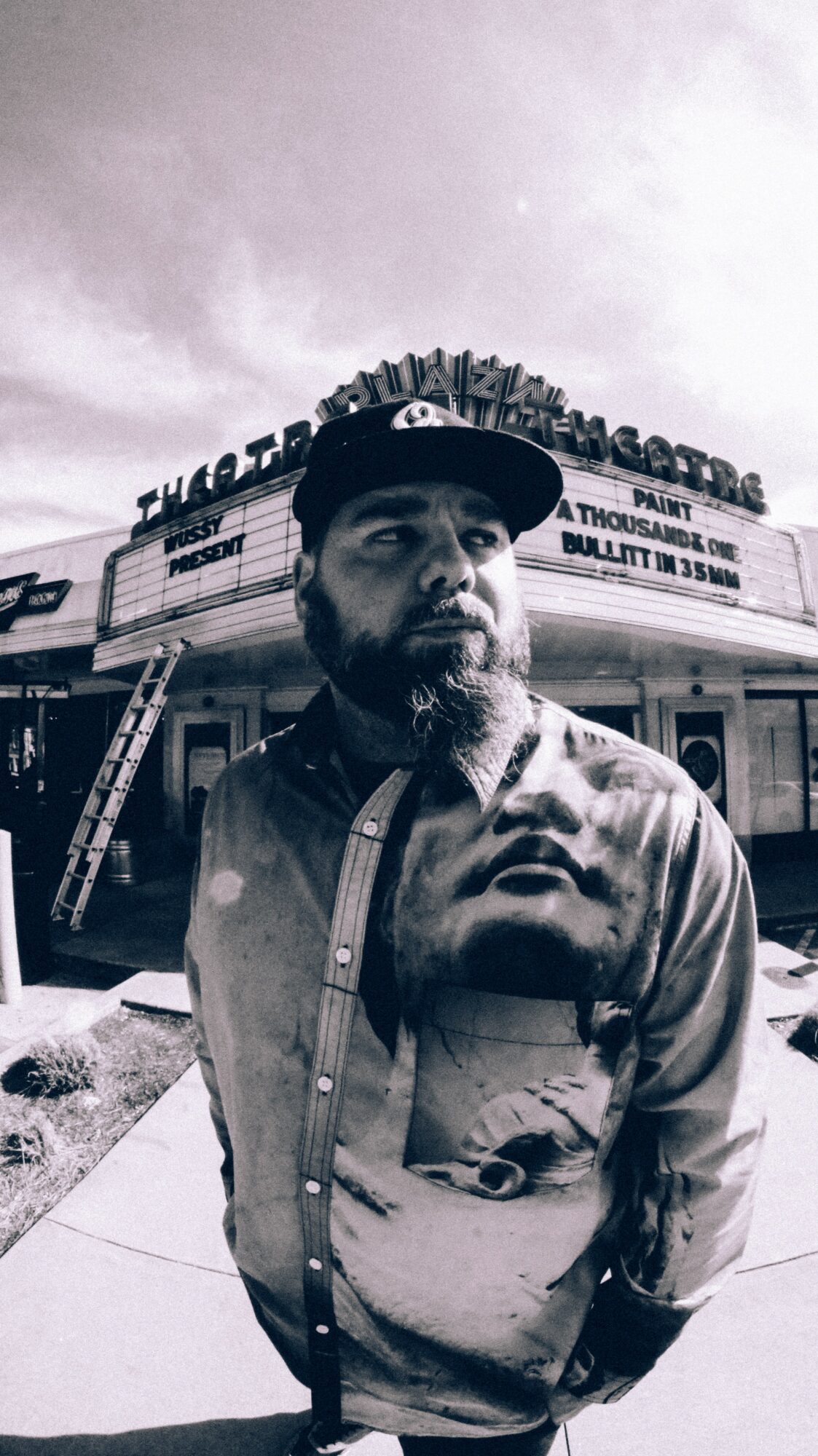 Man with beard and cap standing outside a theater with marquee sign, black and white photo.