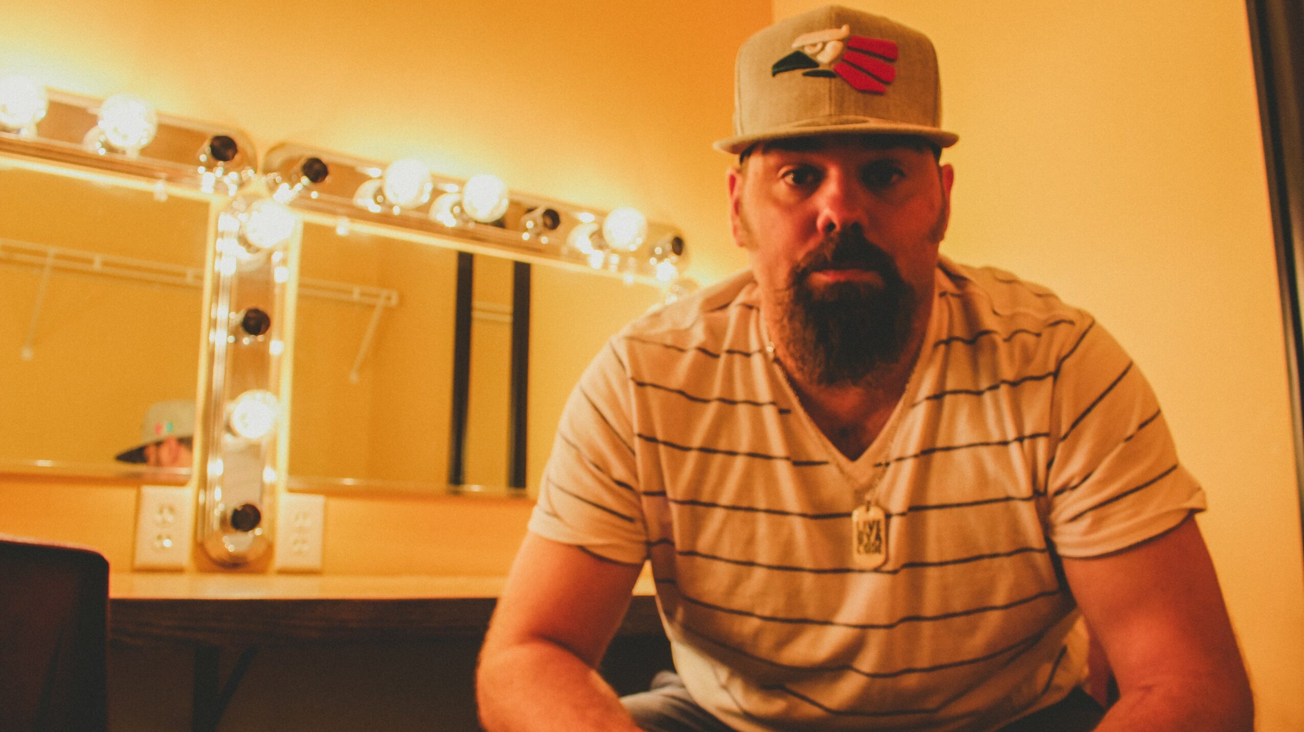 Man with a beard and mustache wearing a striped shirt and baseball cap, sitting in front of a mirror with lights.