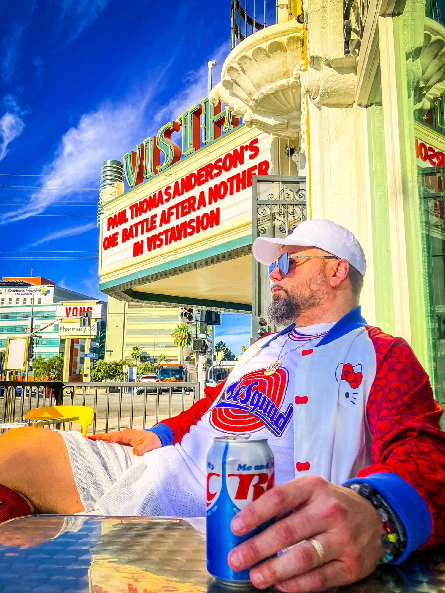 Man with sunglasses and a white cap sitting outdoors at a table, holding a can of soda, with a colorful building and sign in the background.