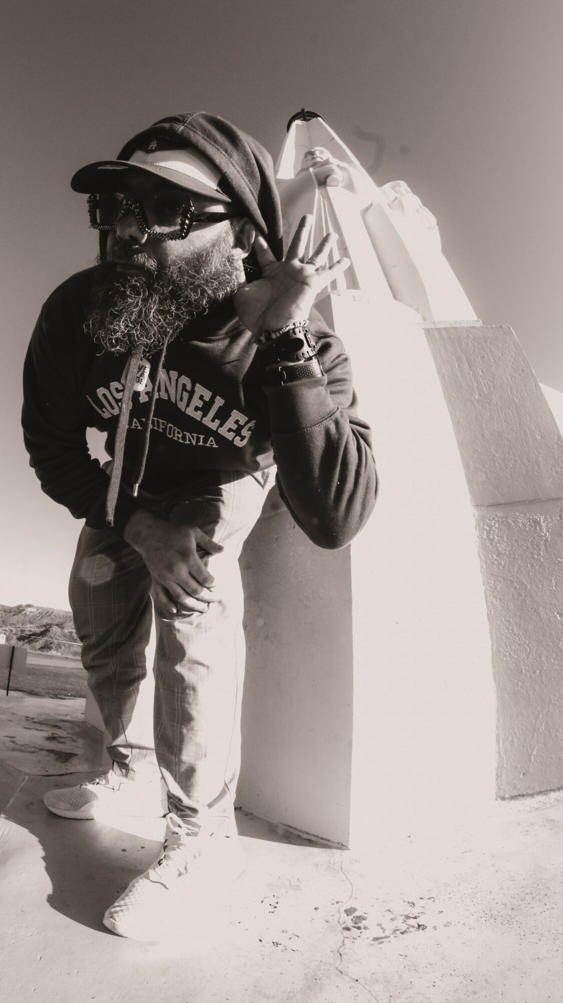 Person with a beard, wearing a cap and sunglasses, leaning against a large structure outdoors, holding a phone to their ear.