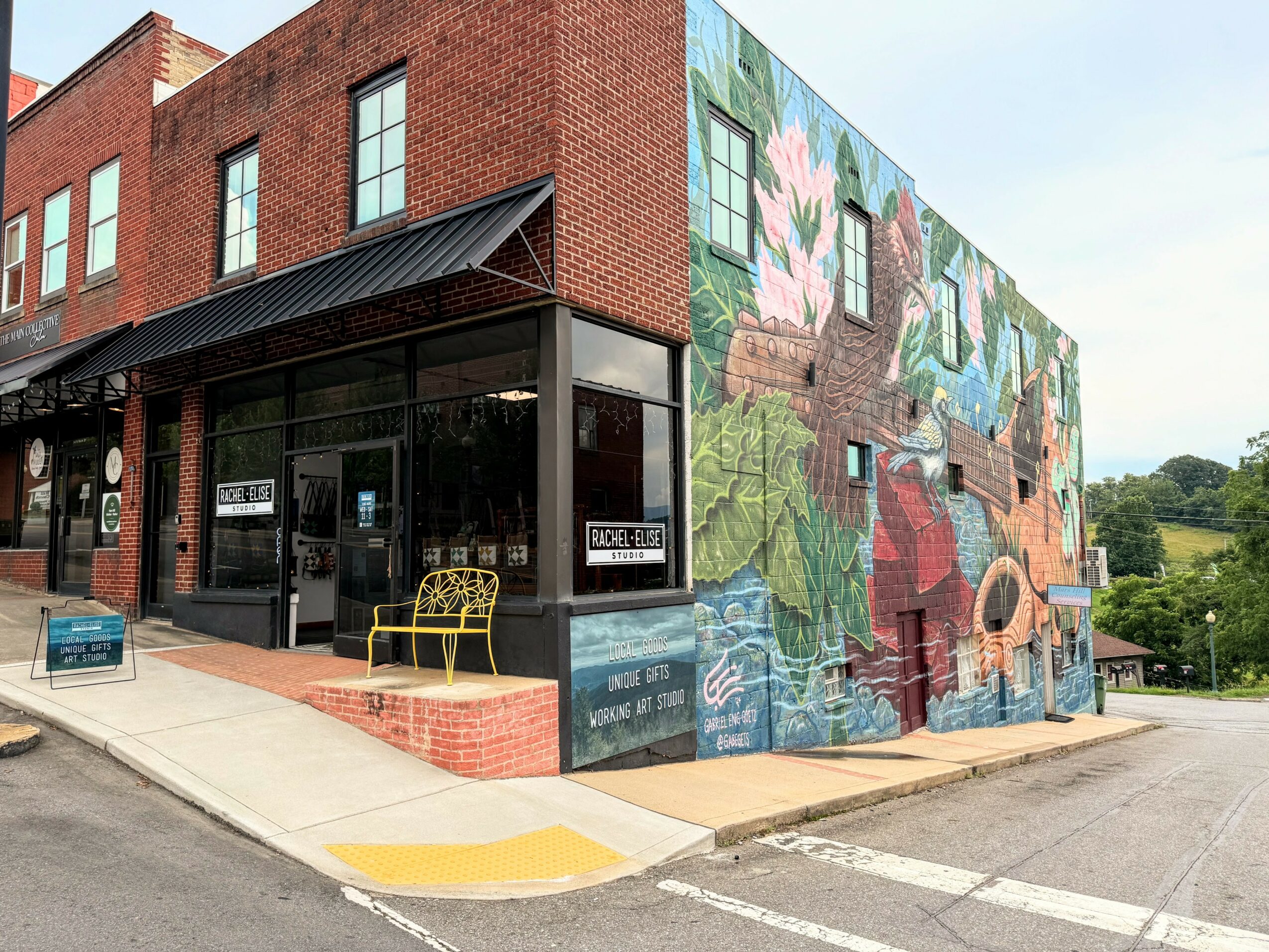 Corner building with a mural of flowers and water, brick facade, glass storefront, yellow bench outside, sidewalk, and street.