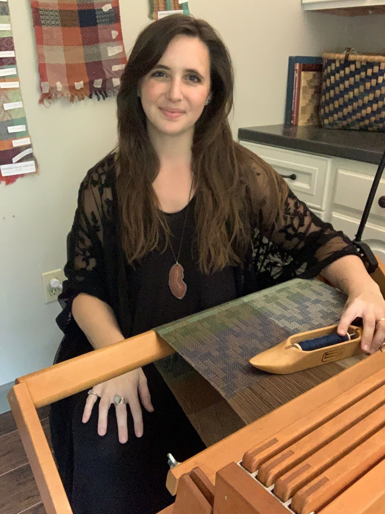 Woman with long dark hair sitting at a table, using a laptop, in a room with a plaid curtain and a cabinet.