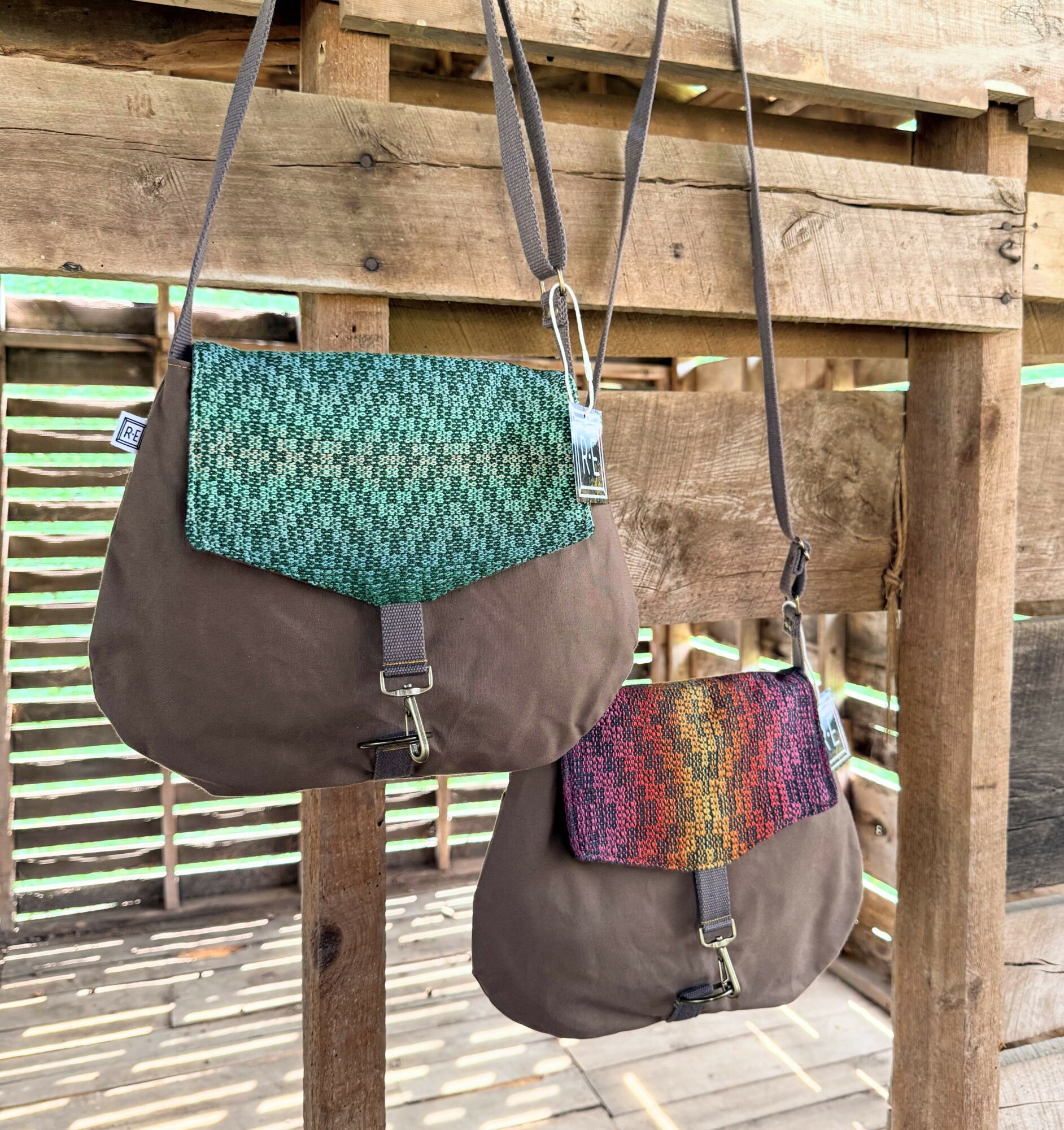 Two brown bags with colorful woven flaps hanging on a wooden fence, sunlight casting shadows. Bag straps hang down.