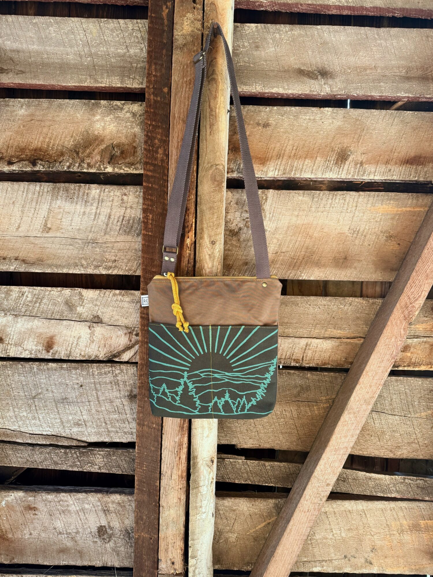 Bag with a landscape design hangs on a wooden fence, strap over a vertical post, against weathered wood background.