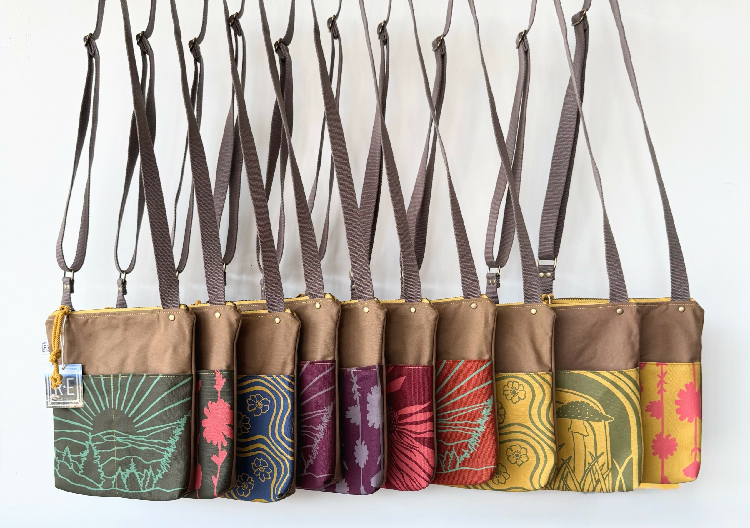 Nine small pouches with straps hanging, each with colorful patterns and designs, arranged in a row against a white background.