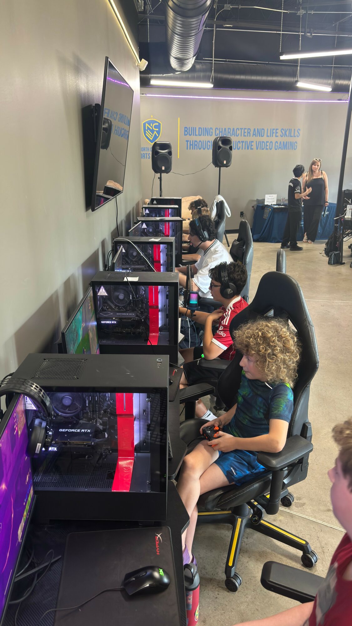 Children seated at computers in a classroom or lab, with adults standing in the background, and a wall with text and equipment.