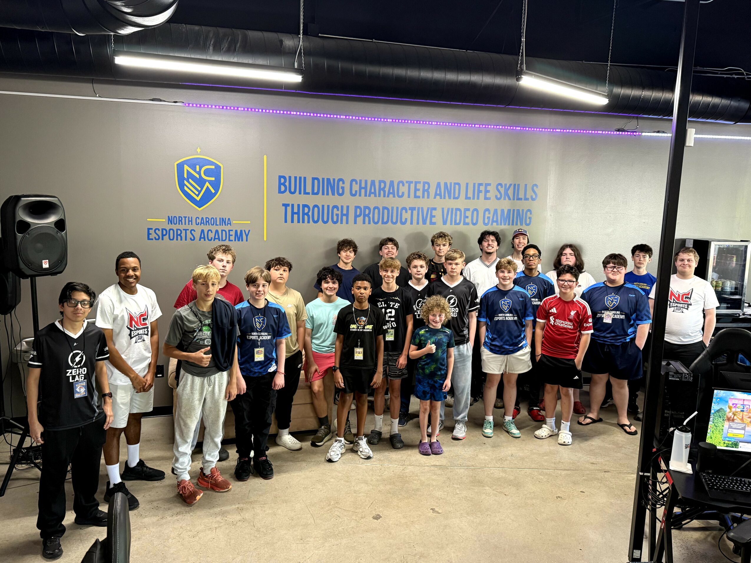 Group of children and adults standing in front of a wall with a North Carolina Esports Academy sign, some smiling.