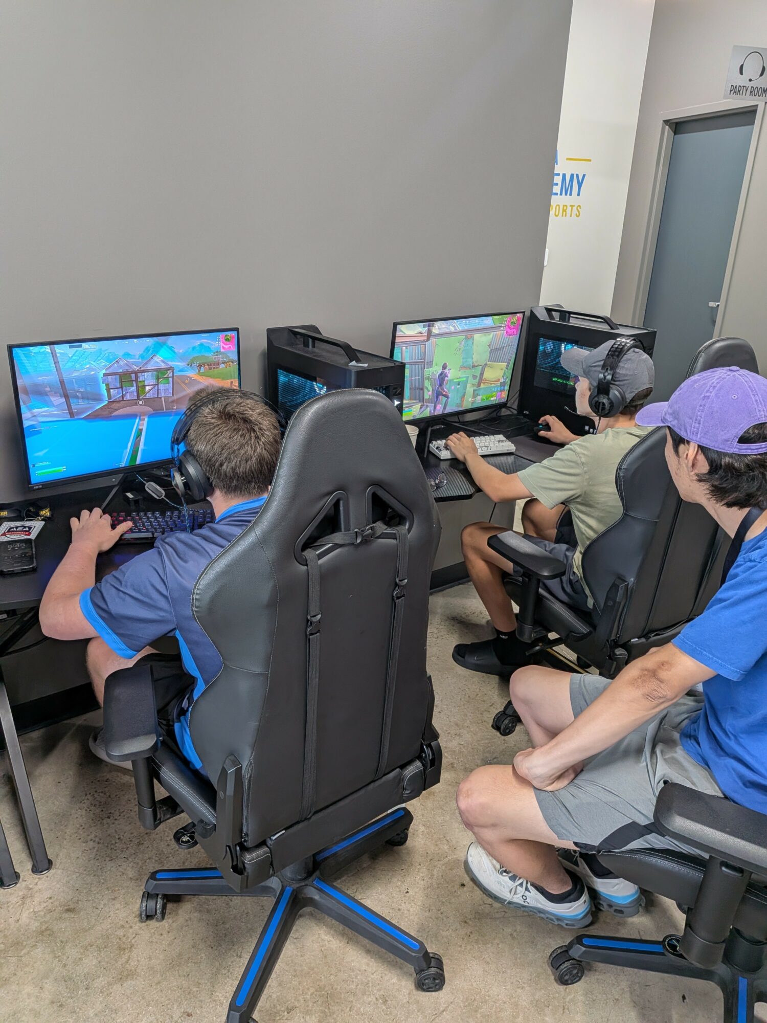 Two children playing video games on computers in a room, one sitting and one standing, with gaming chairs and headsets.