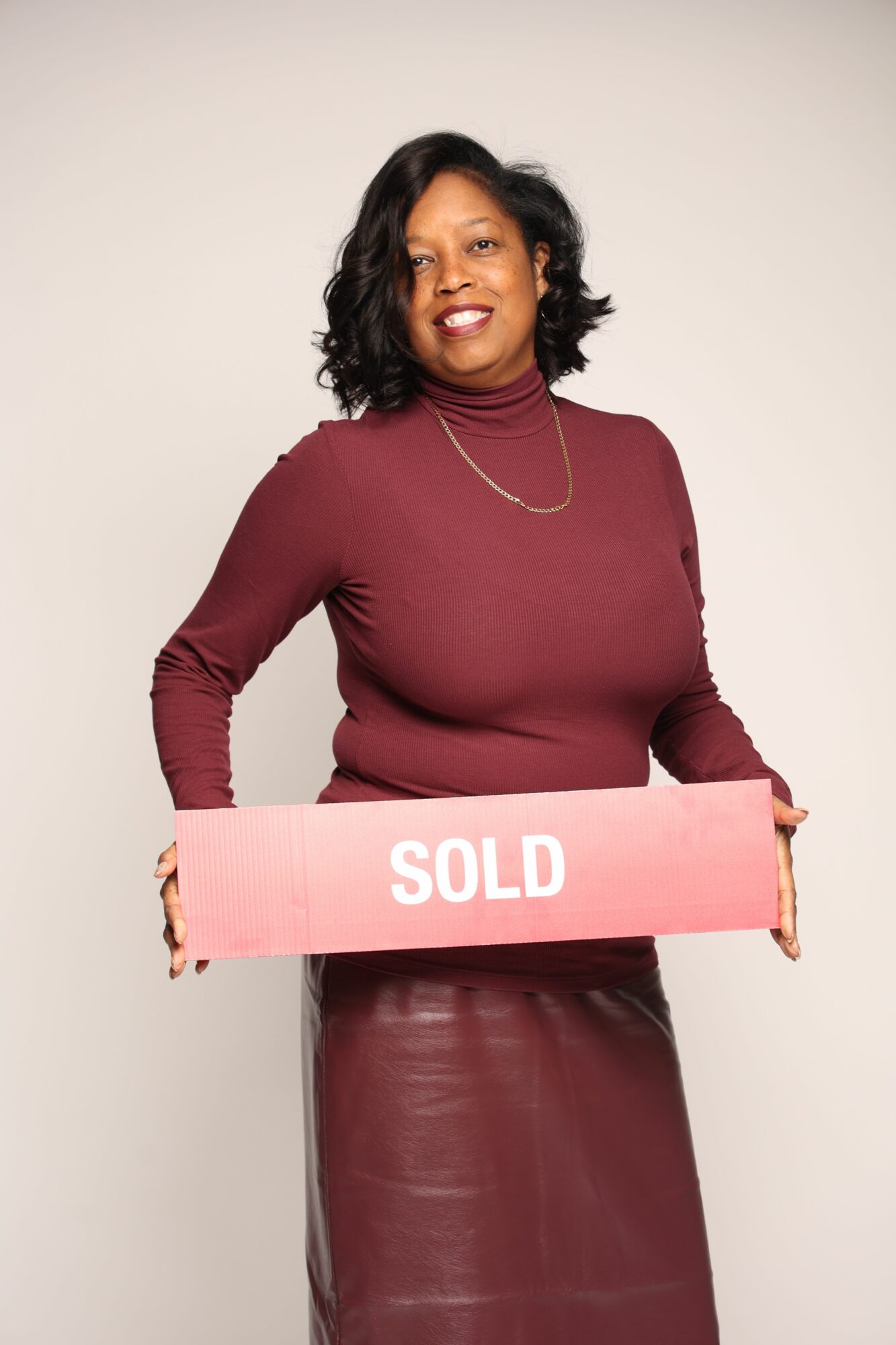 Woman holding a pink sign with the word SOLID, smiling, wearing a maroon dress, standing against a plain background.