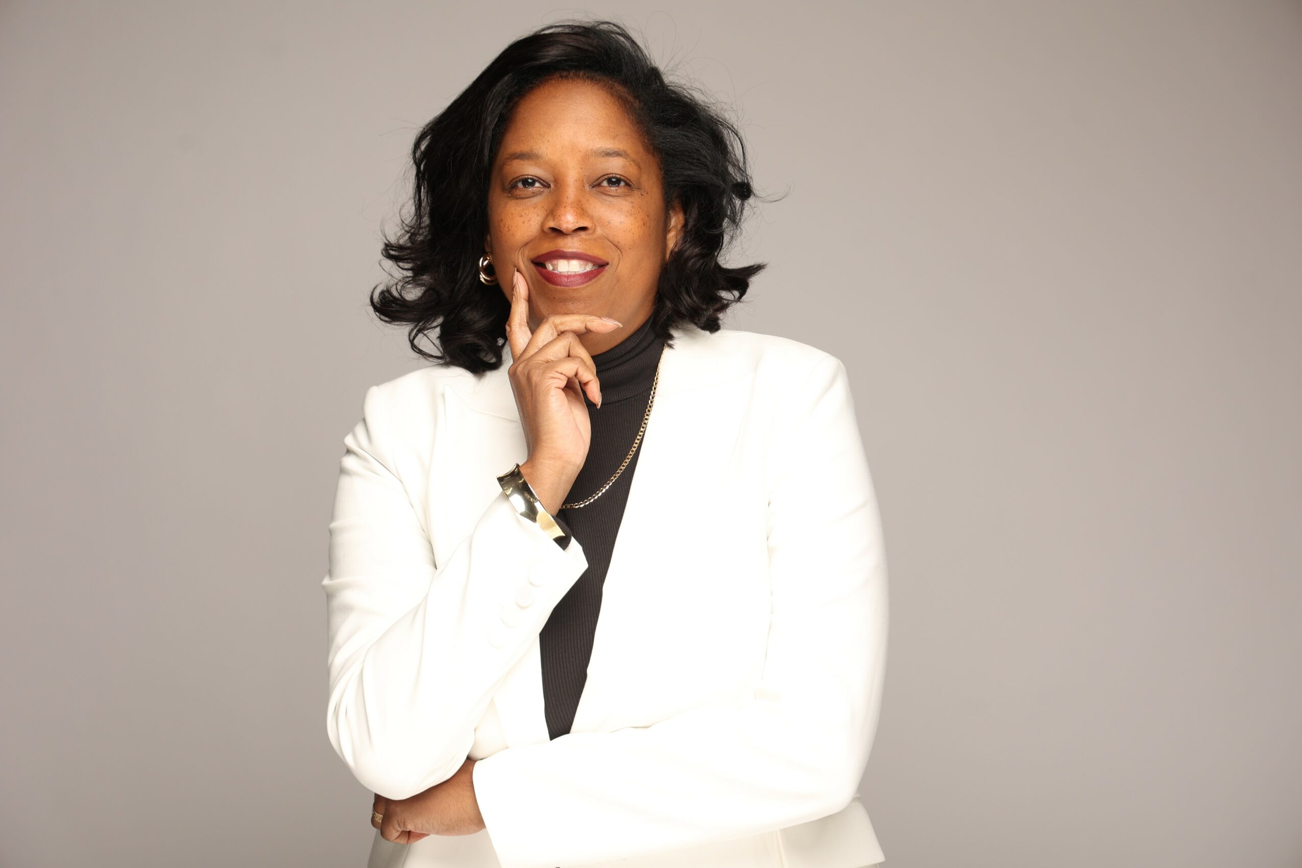 Woman with black hair wearing a white blazer and black top, smiling with hand near chin, neutral background.
