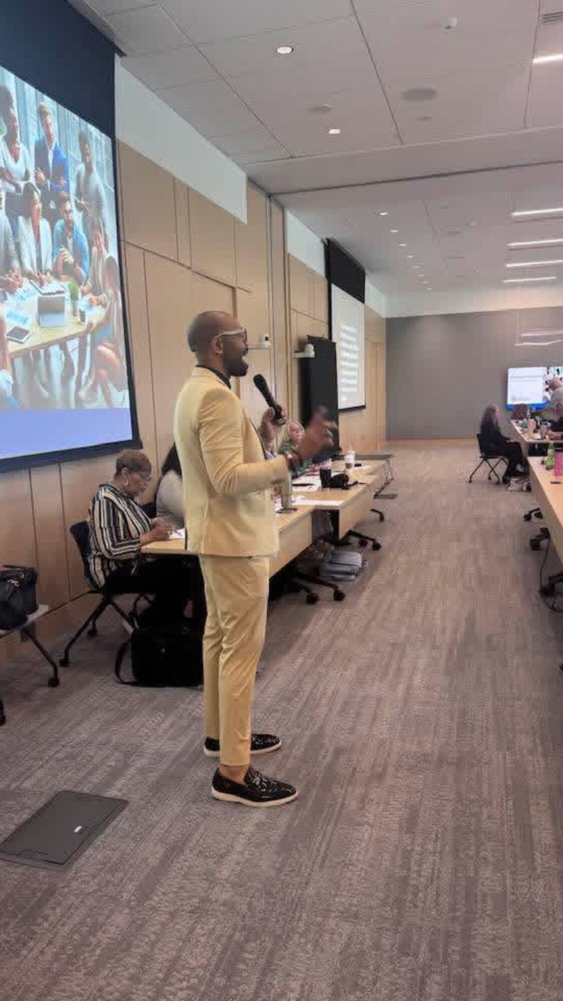 Man in beige suit speaking into microphone in conference room with large screen and seated audience.