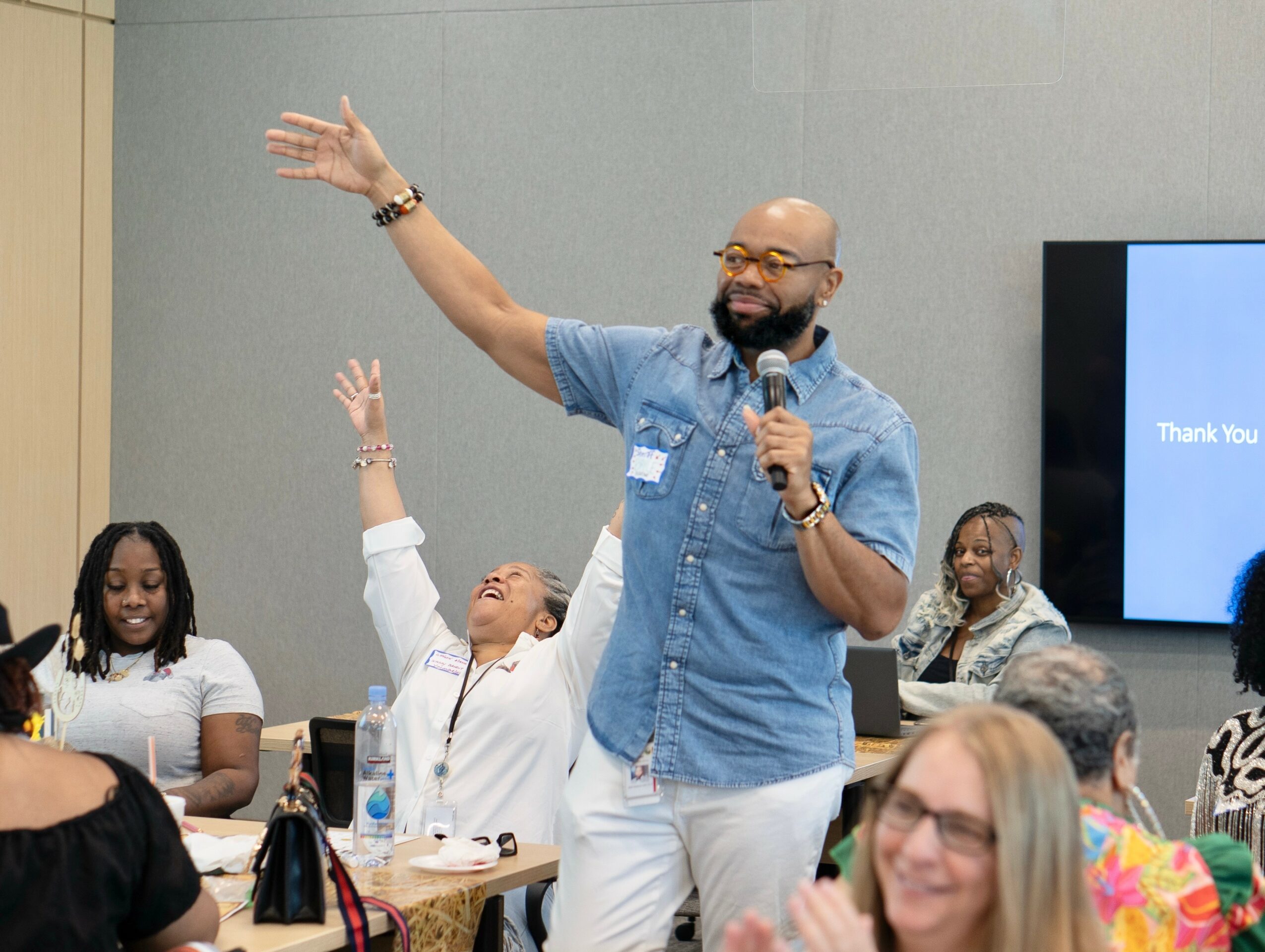Man speaking into microphone with audience raising hands, seated at tables, in a conference room.