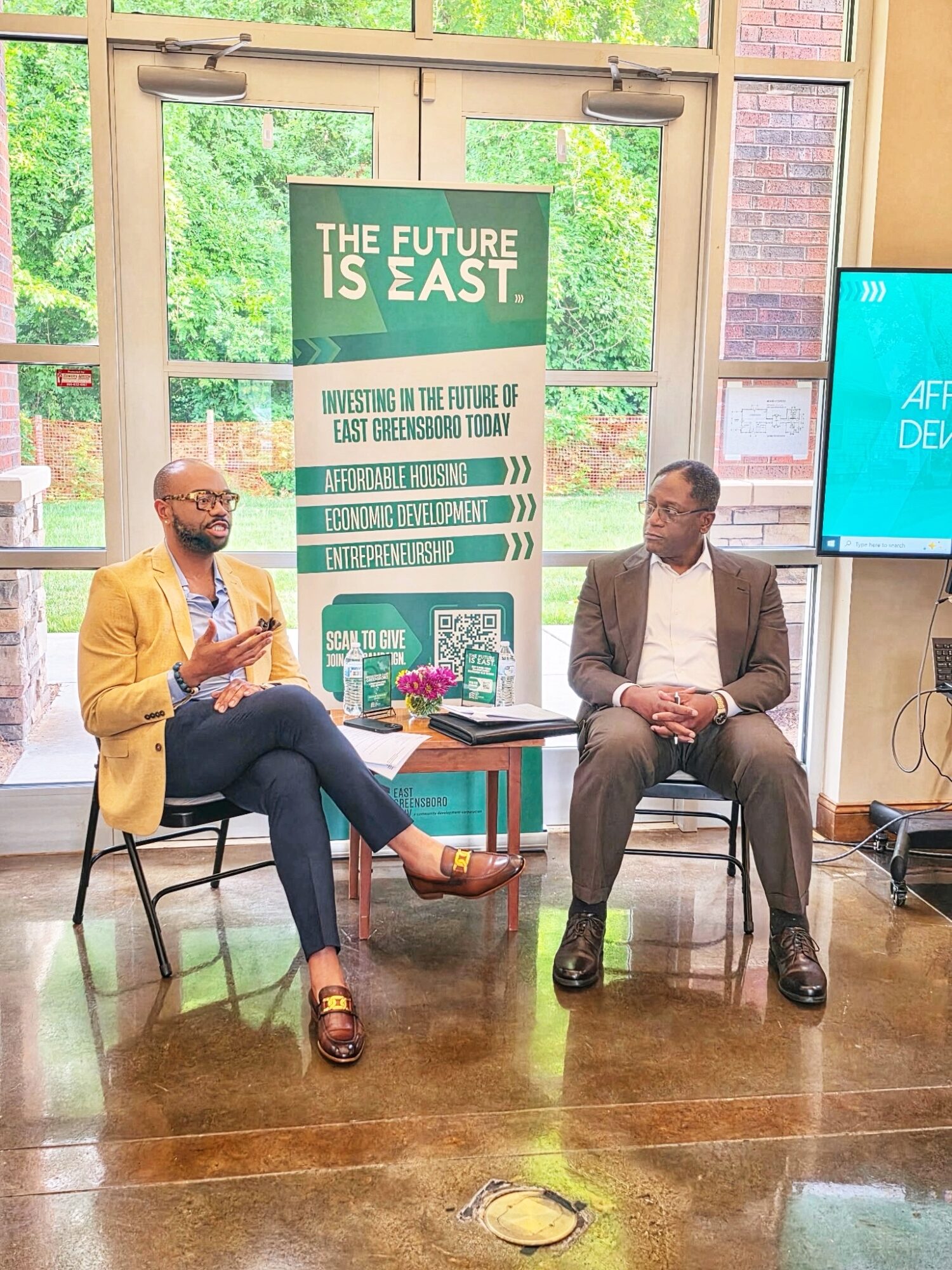 Two men seated in a room with large windows, discussing at a panel event, with a green poster behind them.