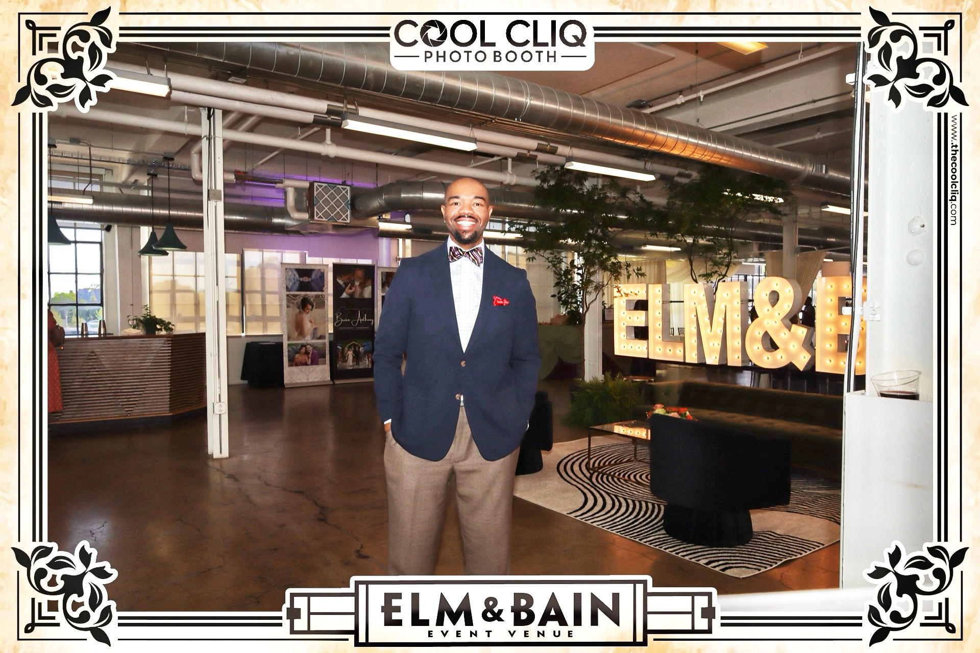 Man in a suit and bow tie standing inside a photo booth with illuminated 'ELM & BAIN' sign behind him.