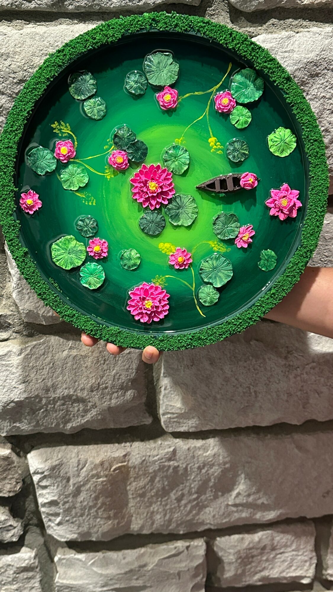Round decorative water feature with pink flowers, green lily pads, a fish, and a small frog, held against a stone wall.