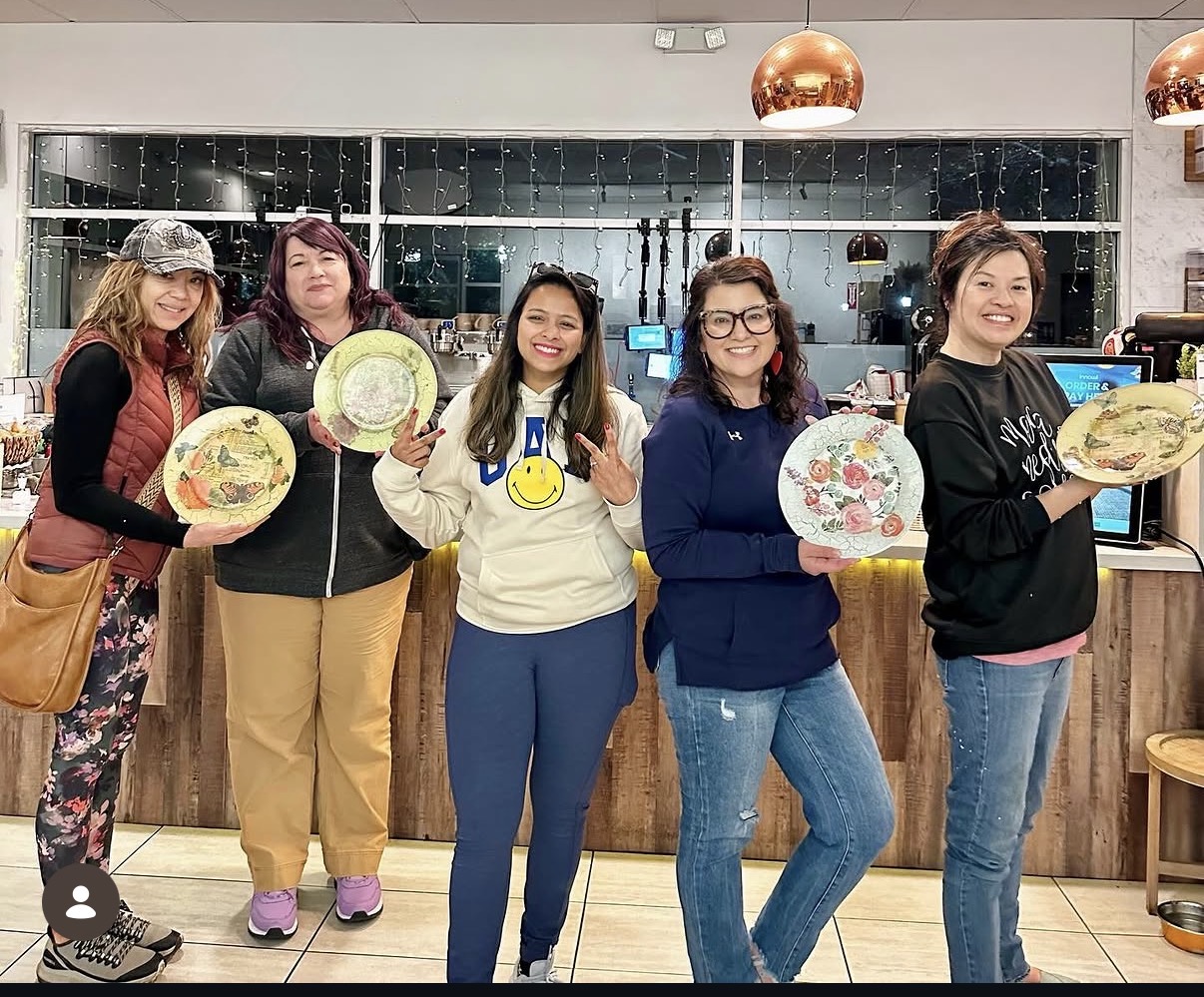 Five women standing in a row holding decorative plates, smiling, in a modern indoor setting with a glass wall and wooden counter.