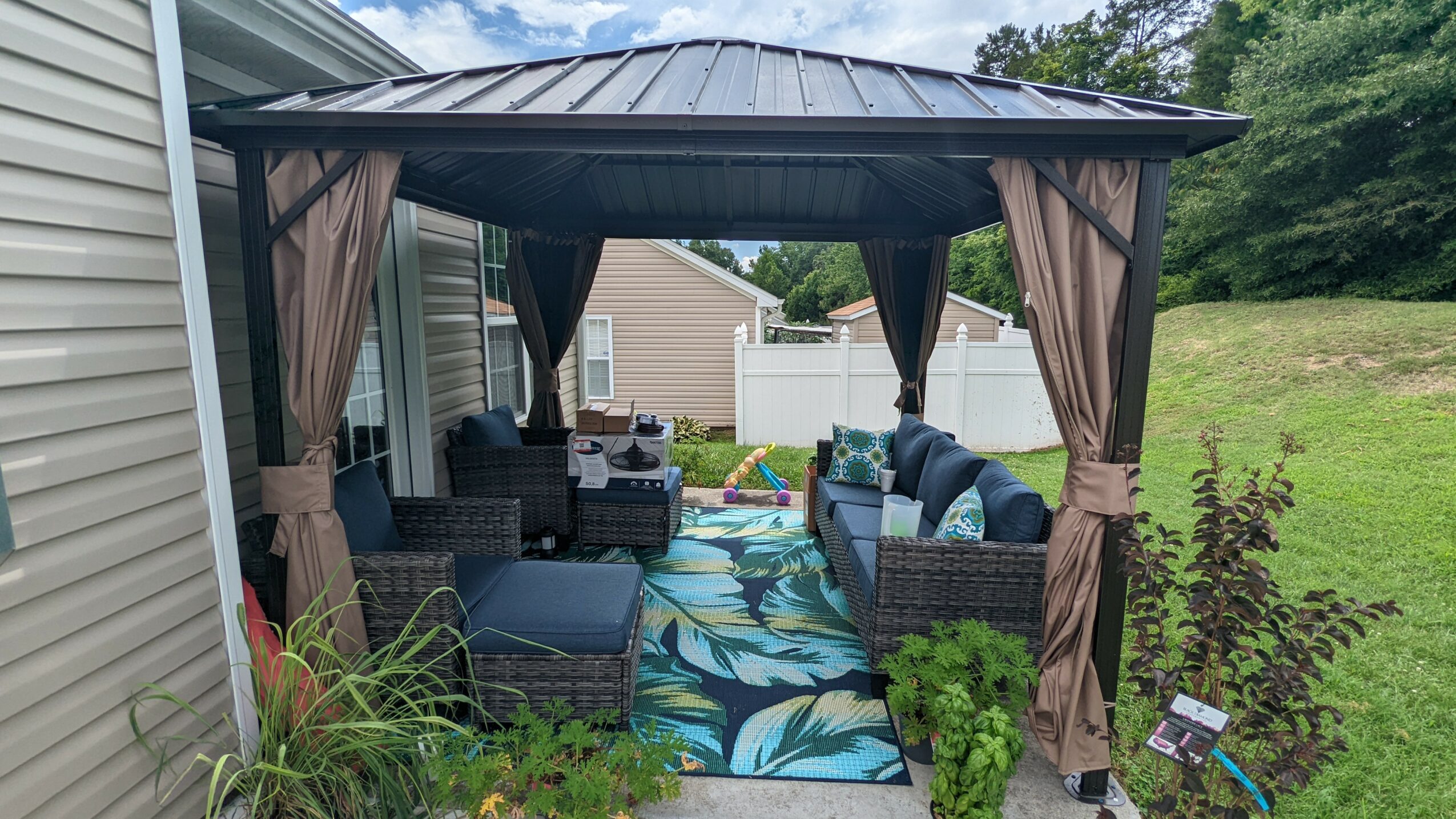Outdoor gazebo with seating area, curtains, and a colorful rug, surrounded by greenery and a house in the background.