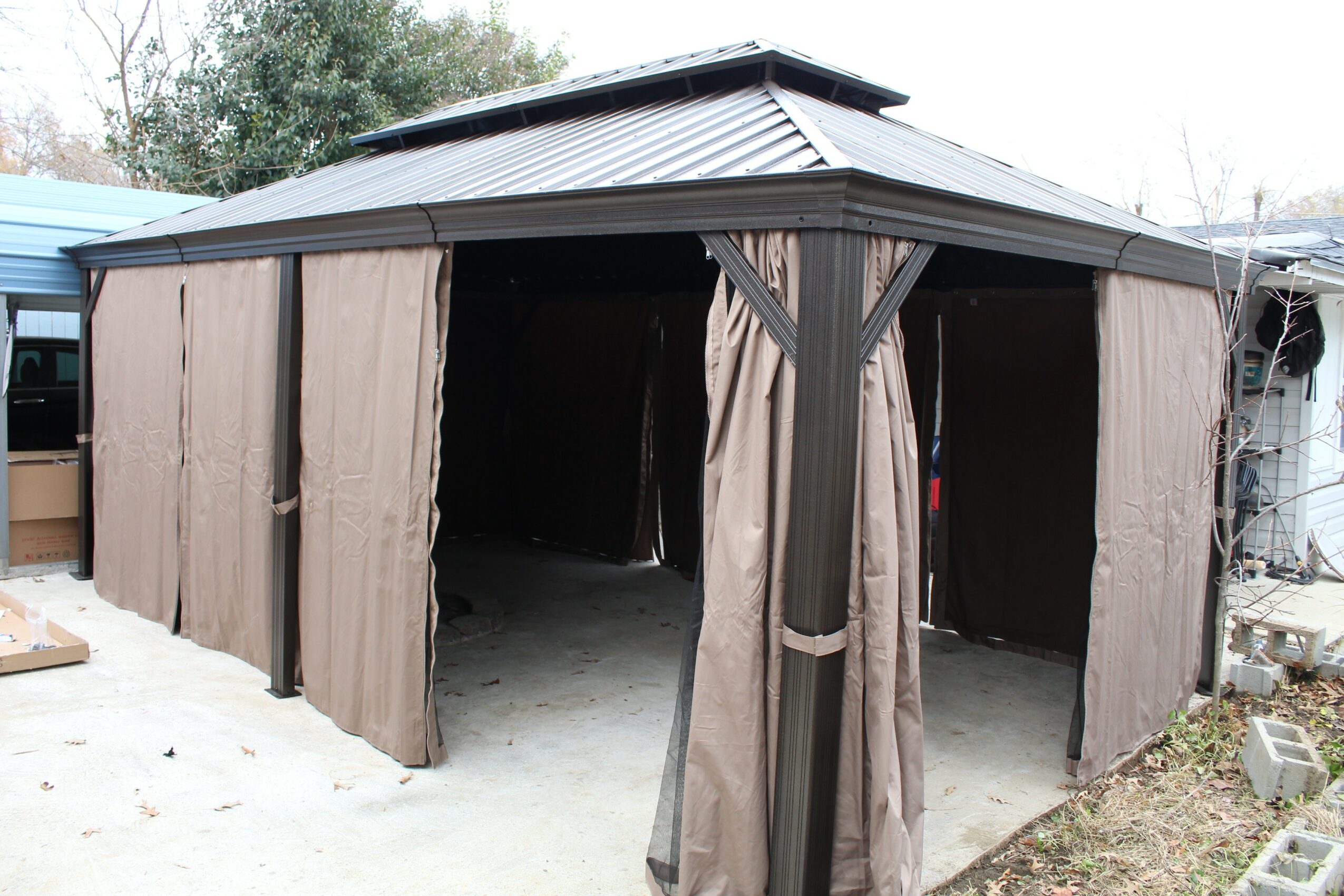 Outdoor gazebo with brown curtains, open sides, and a metal roof, surrounded by trees and grass.