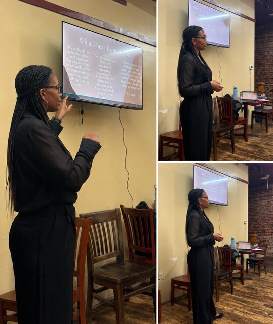 Woman giving a presentation in front of a screen, with chairs and a table in the background.