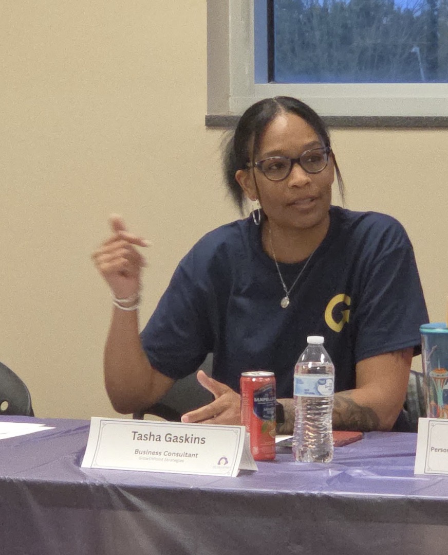 Woman with glasses and dark hair, wearing a navy shirt with a yellow letter G, sitting at a table with drinks and a nameplate.